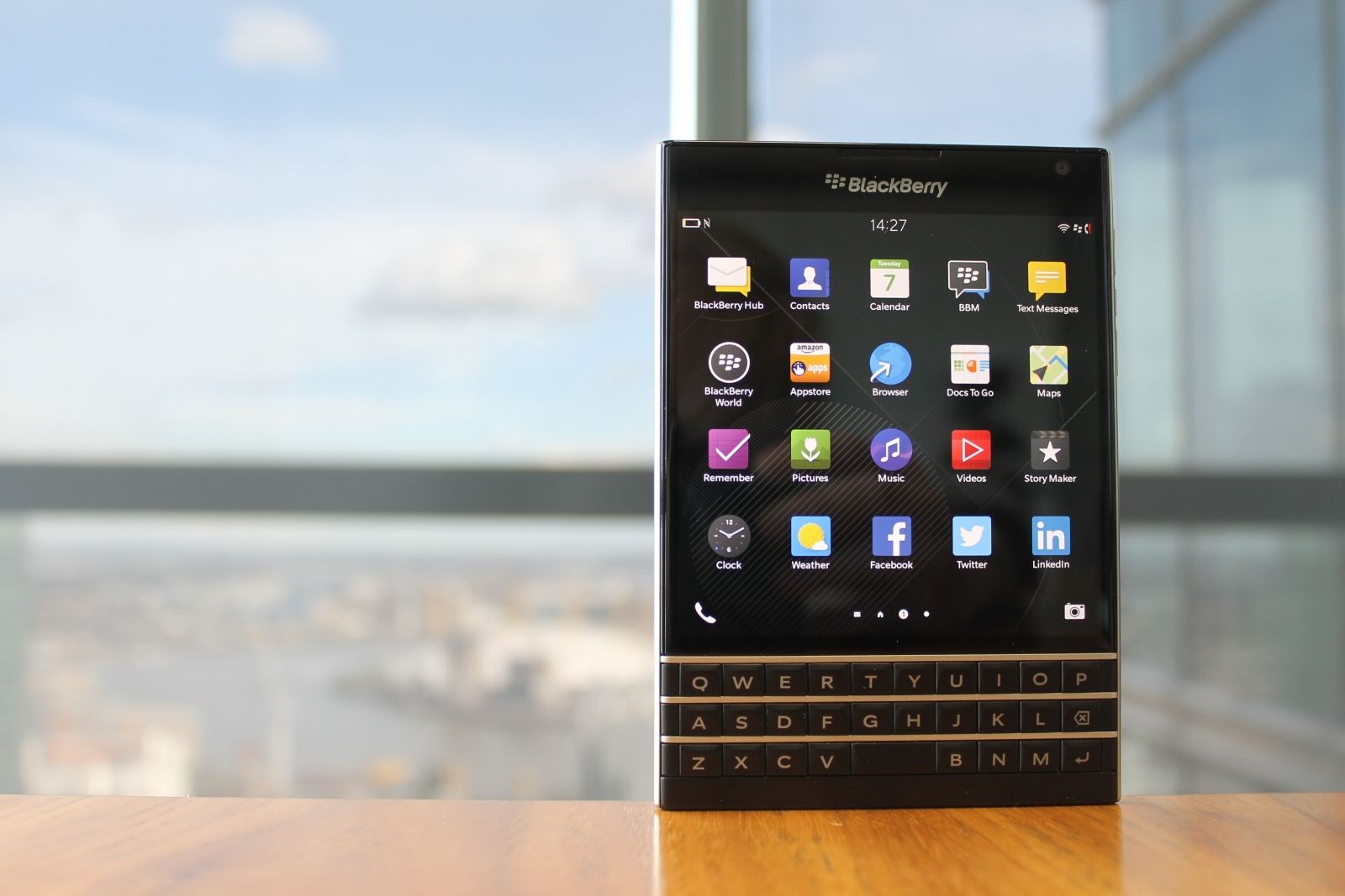 BlackBerry telefon fa asztalon. Alkalmazások a képernyőn, billentyűzet alul. Épületek és égbolt a háttérben.