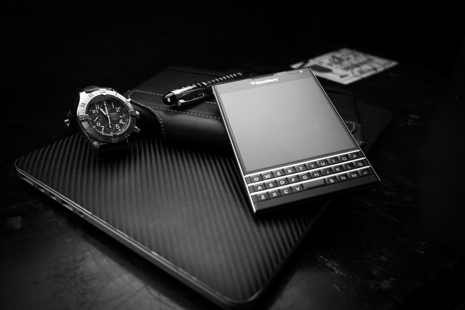 Egy óra, egy toll, egy Blackberry telefon és egy jegyzetfüzet szénszálas felületen. A jelenet fekete-fehér.