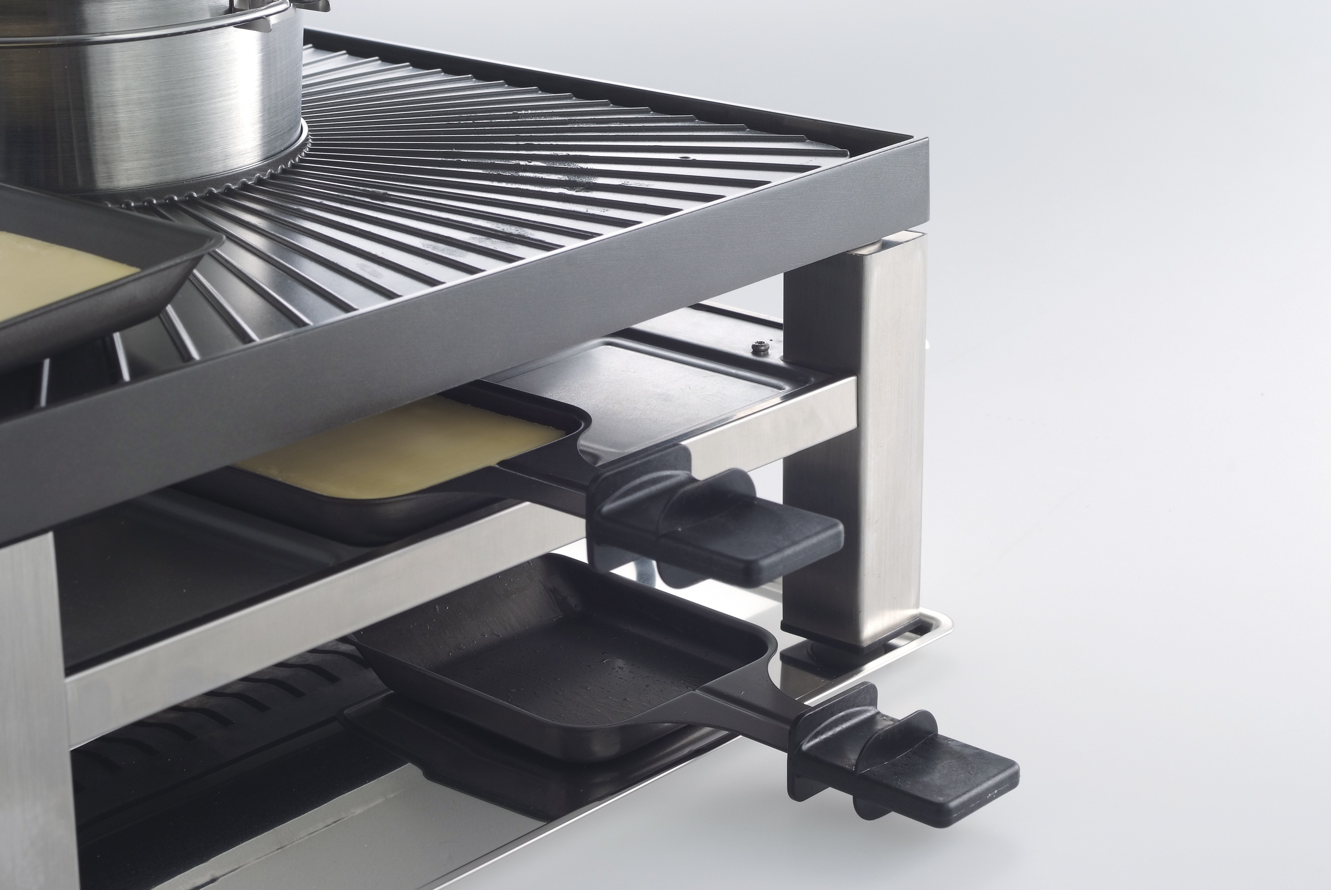 Raclette grill with trays and heating elements visible. Stainless steel and black materials. White background.