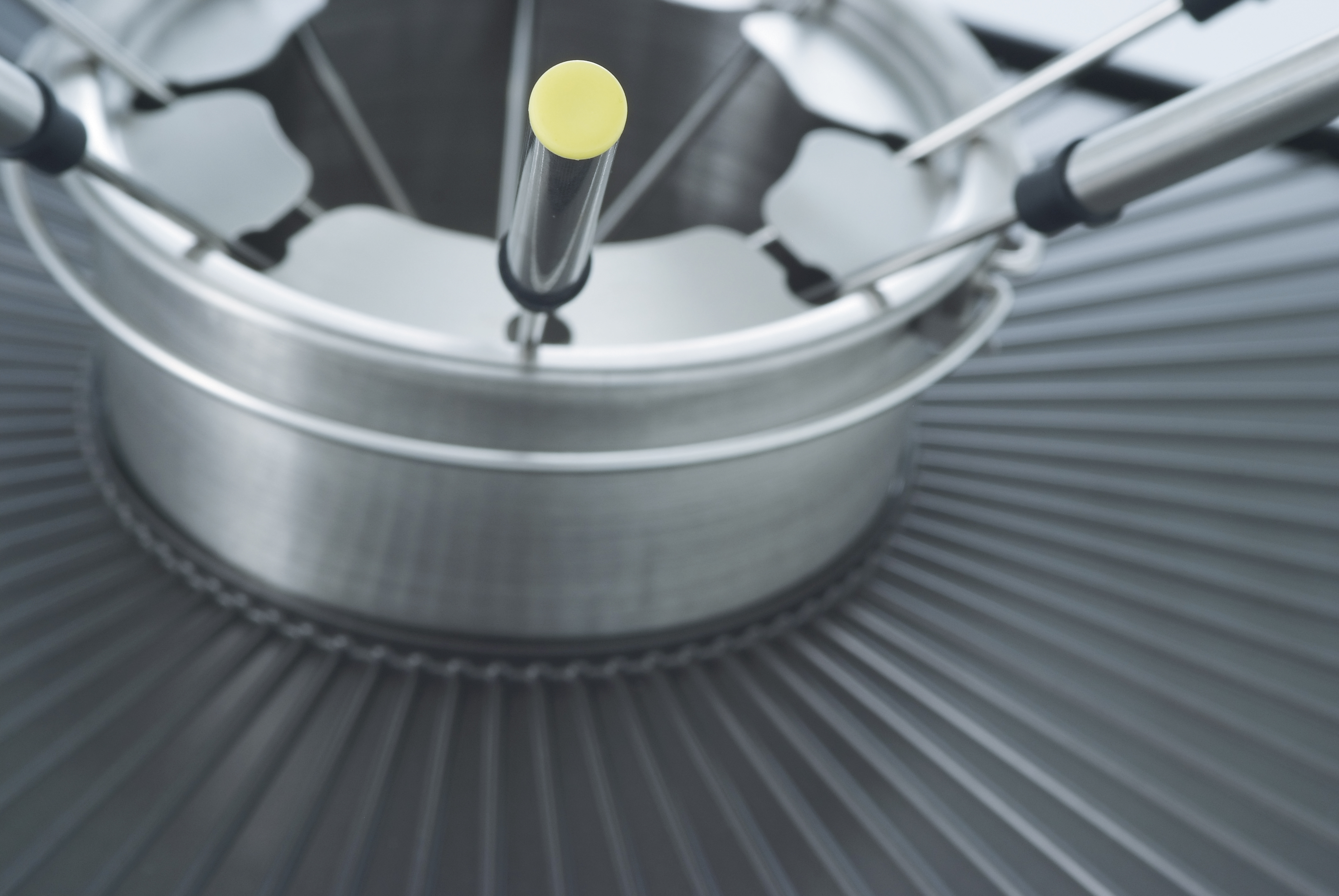 A stainless steel fondue set with a yellow-tipped handle in the center, on a metal grate surface.