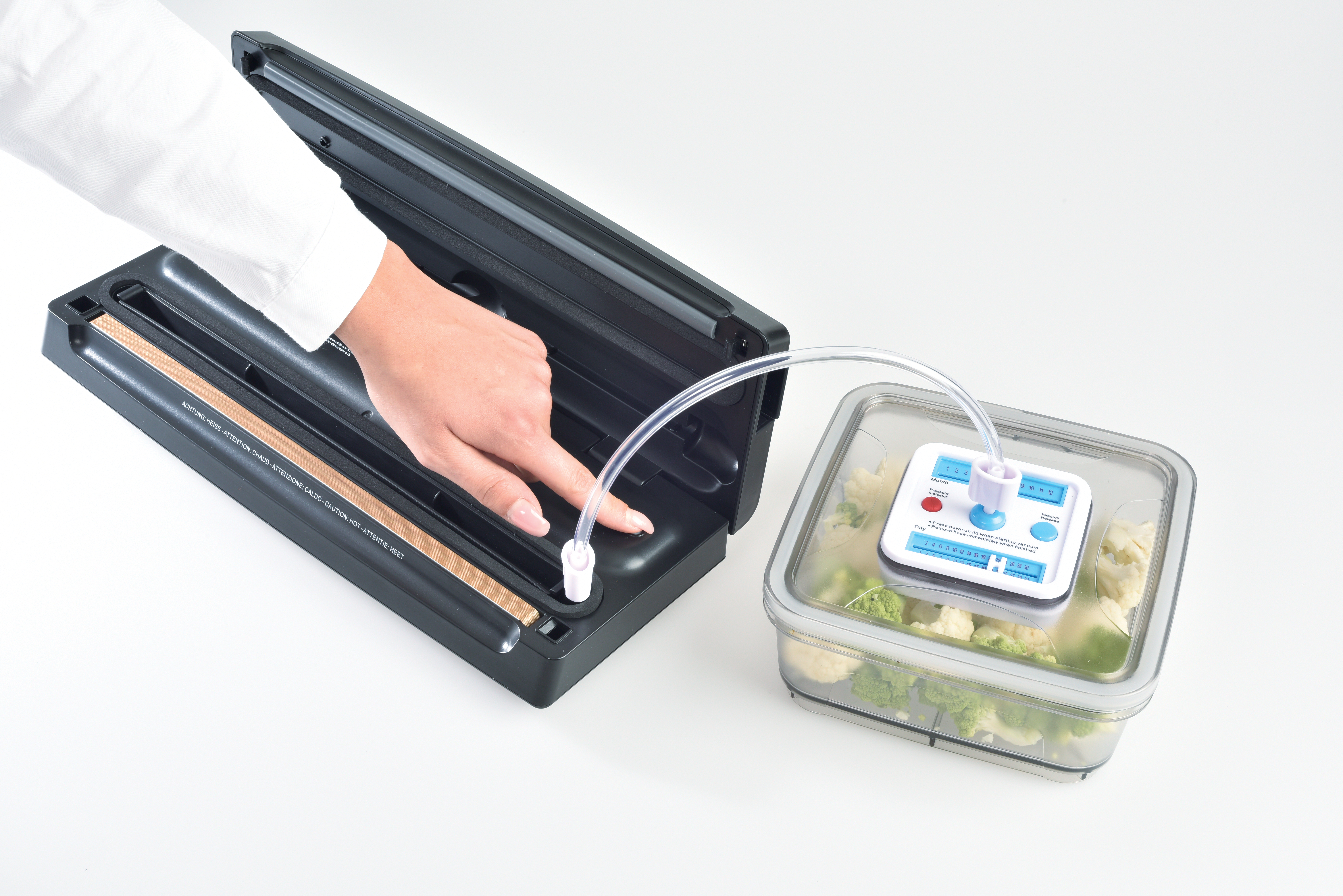 A person operates a vacuum sealer with a hand. A container with vegetables is connected via a tube. The machine is black.