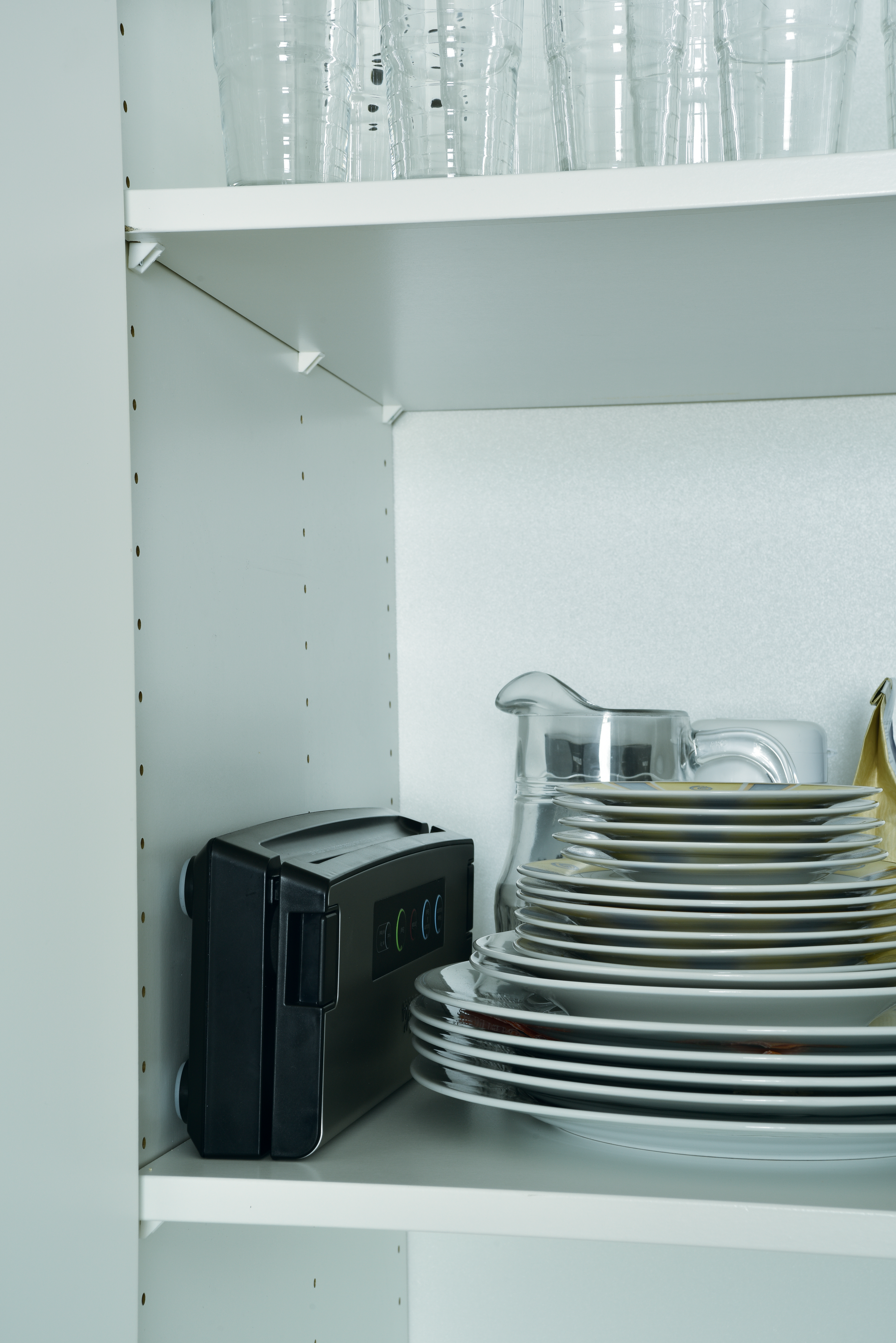 Inside a white cabinet, there are glasses, a black device, plates, and a pitcher. The cabinet is white with multiple shelves.