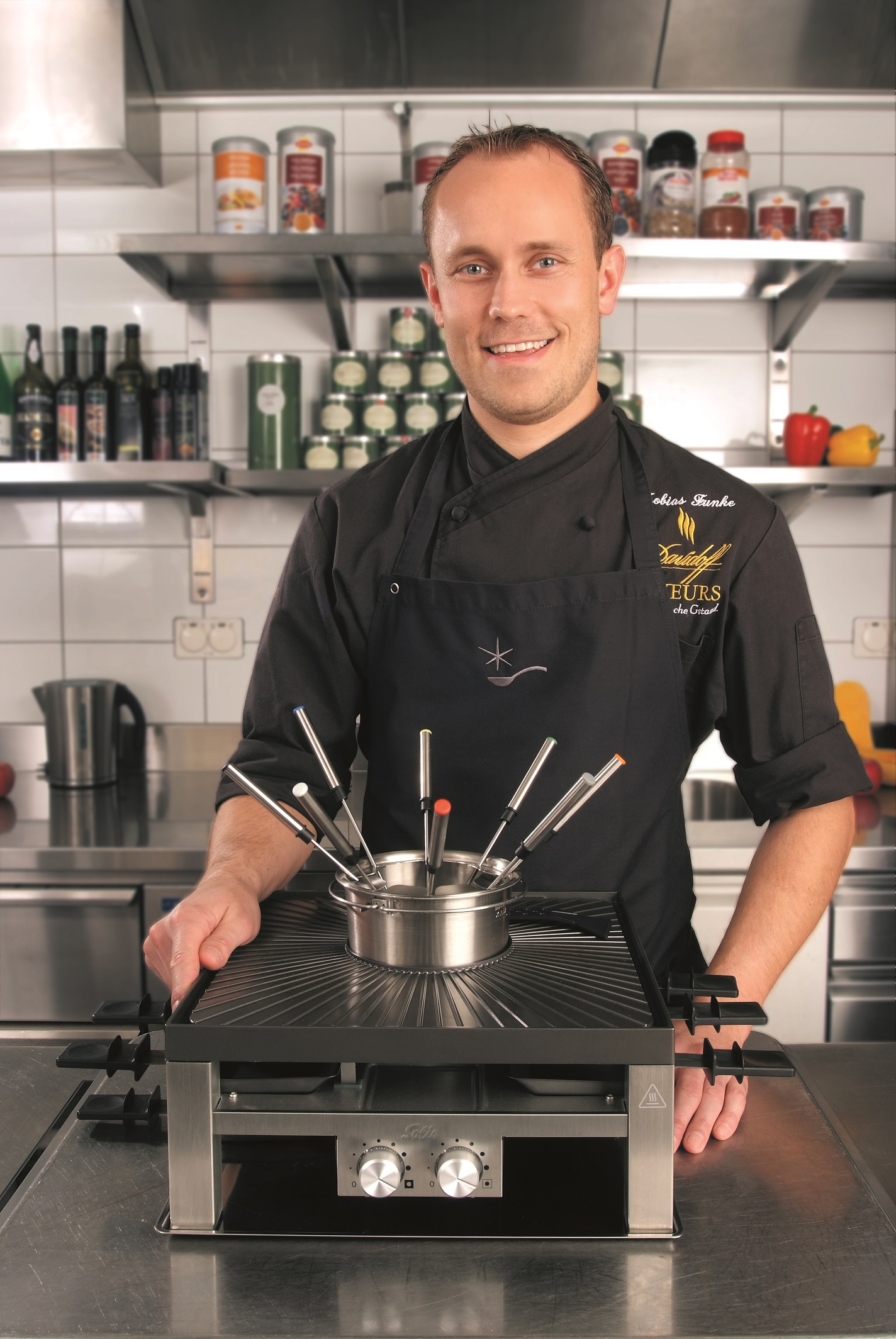 Chef smiling, holding fondue pot over a grill. Kitchen background. Various utensils.