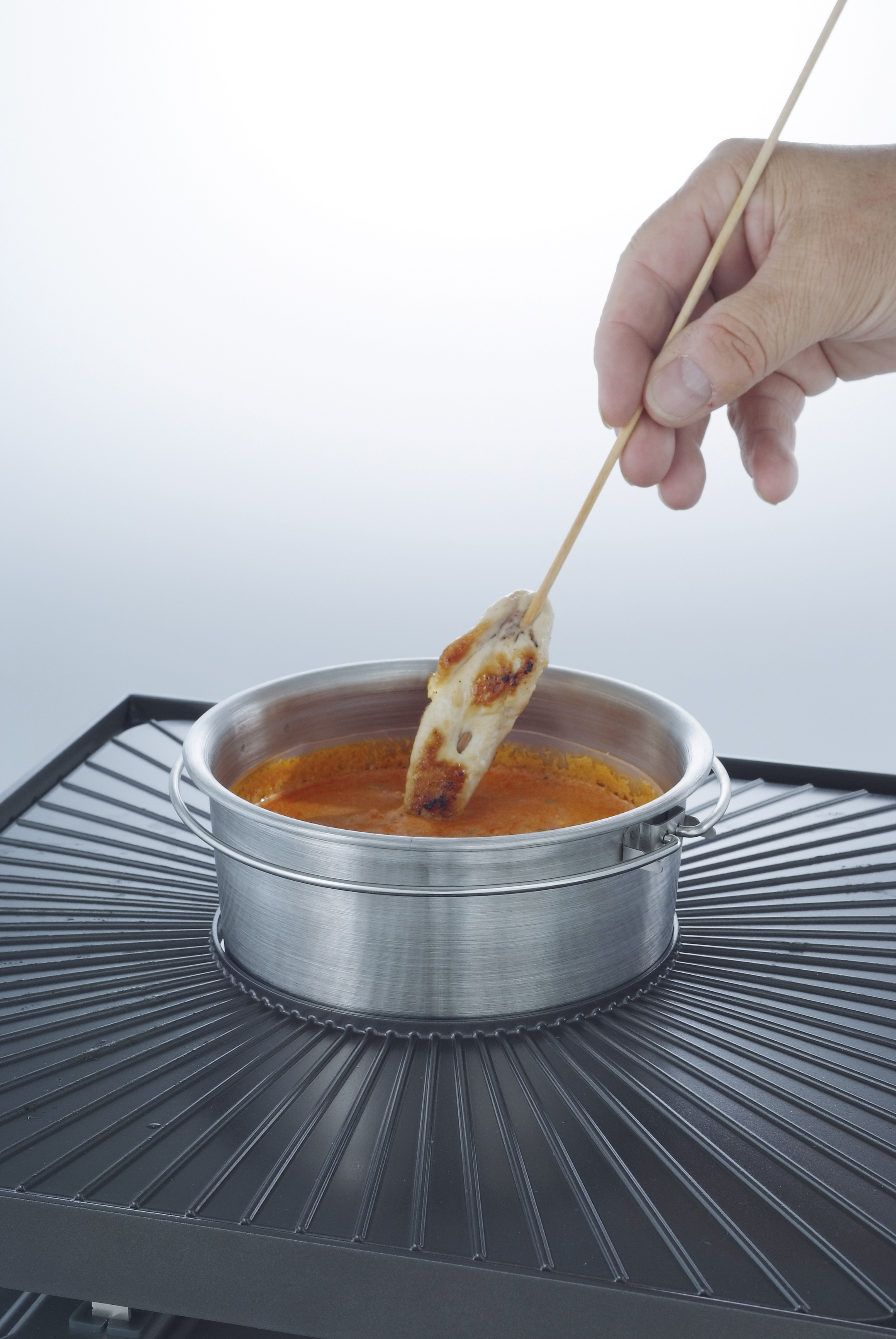 A hand dips a skewer with food into a bowl of sauce on a grill.