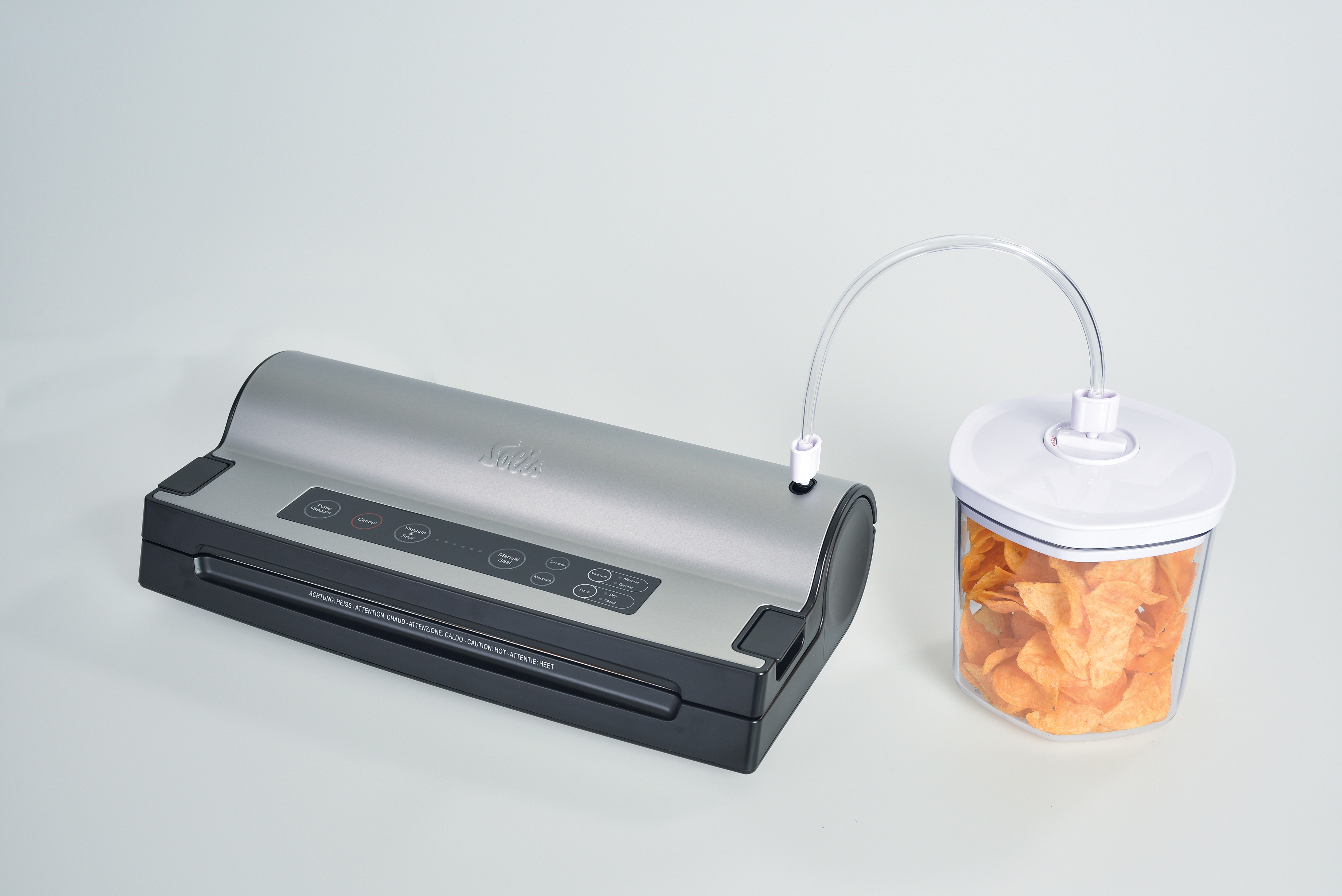 Een zilveren en zwarte vacuümverpakker is via een doorzichtige buis verbonden met een doorzichtige bak met chips.