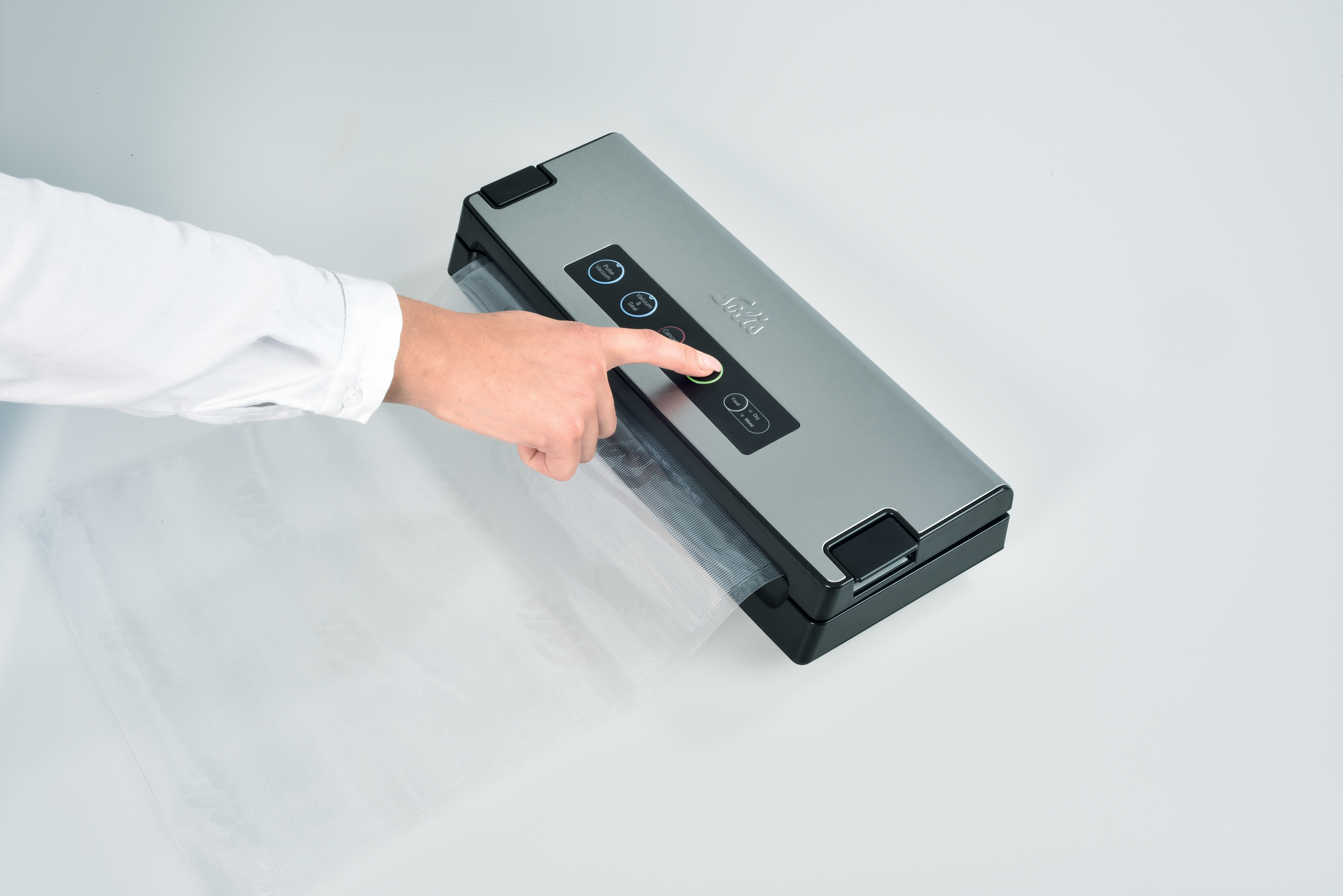 A person's hand presses a button on a silver vacuum sealer. A clear plastic bag is being sealed. White background.