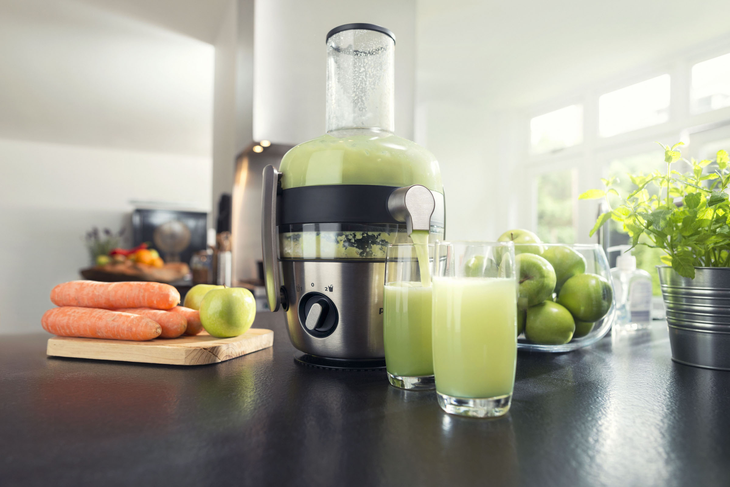Entsafter mit grünem Saft, der gegossen wird. Karotten, grüne Äpfel und eine Pflanze auf einer schwarzen Arbeitsplatte.