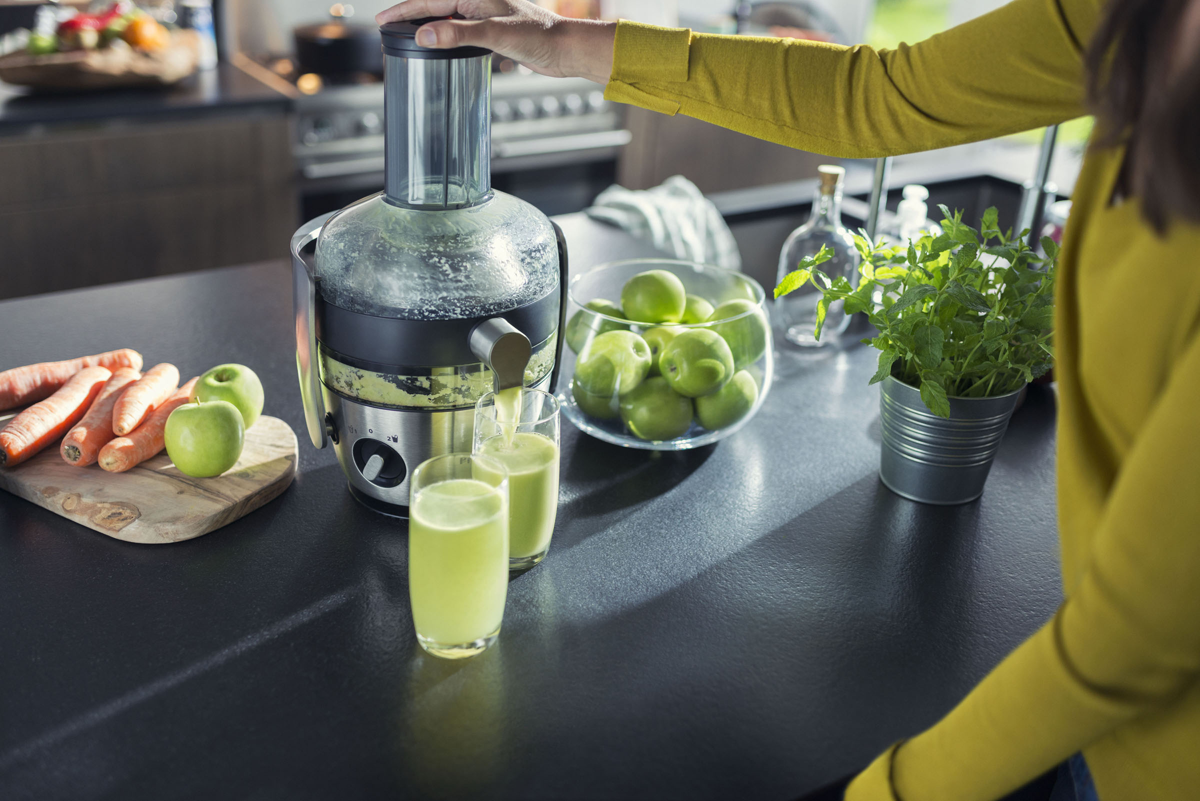 Person gießt Saft aus einem Entsafter in Gläser, mit Karotten, Äpfeln und Pflanzen in der Nähe.