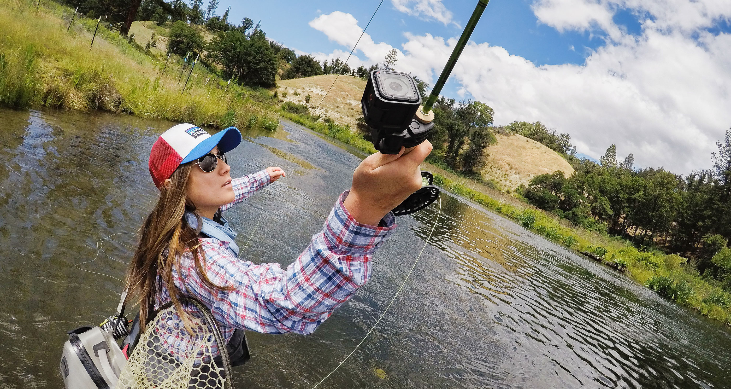 Woman fly fishing in a river. Holding a GoPro camera on a rod. Scenic landscape with mountains and trees.