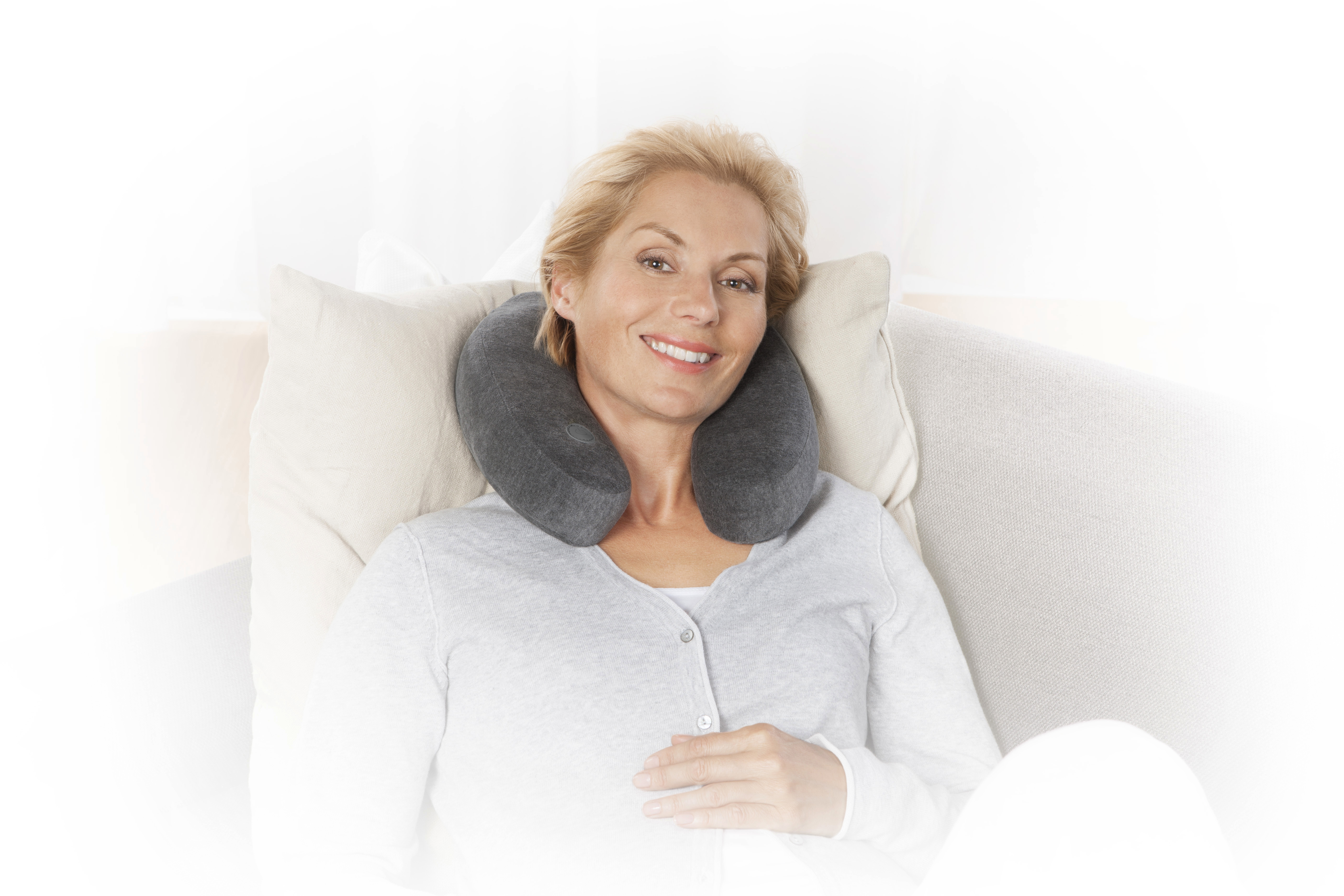 Eine blonde Frau liegt auf einem Sofa mit einem grauen Nackenkissen. Sie lächelt und trägt eine hellgraue Strickjacke.
