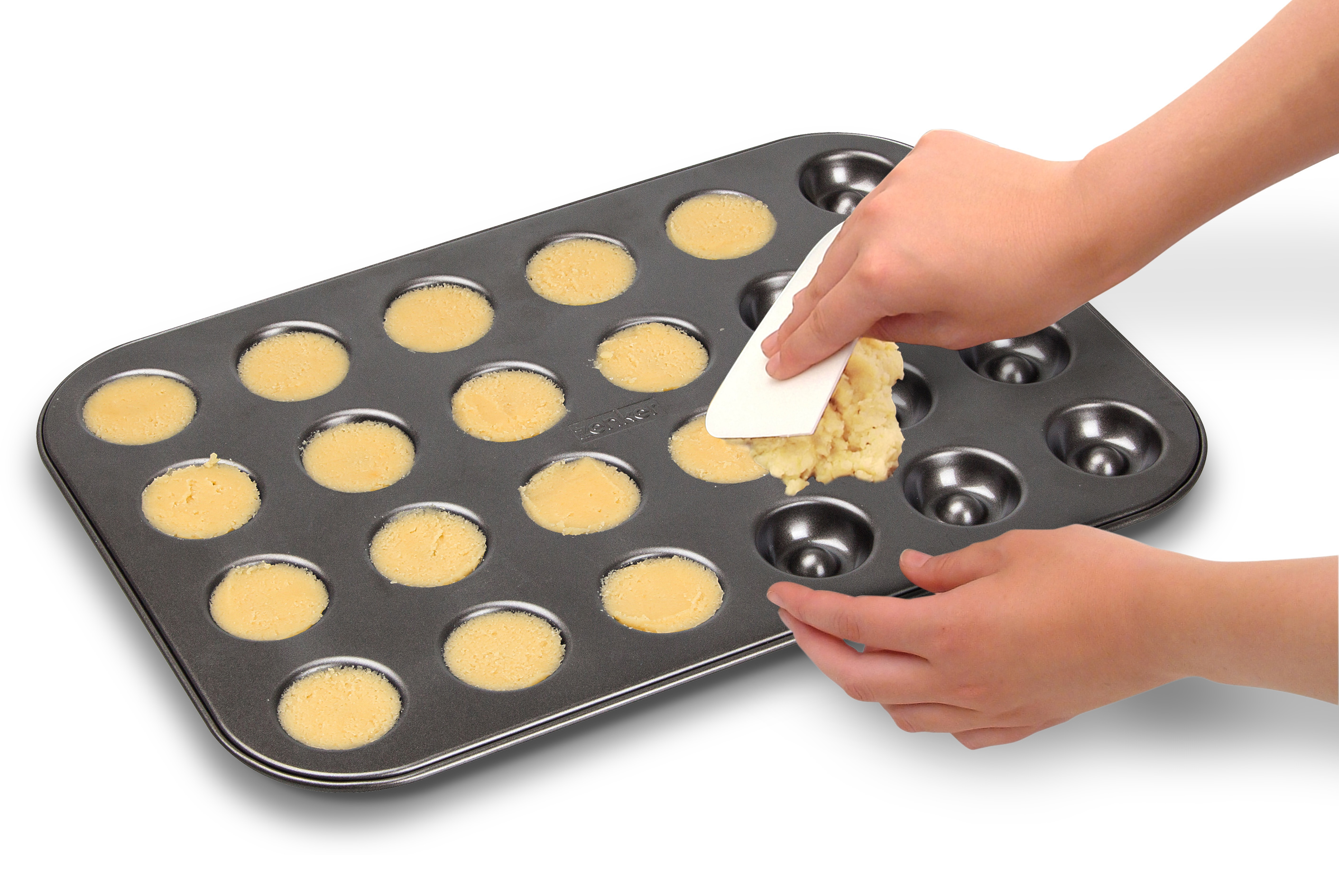Hände einer Person, die einen Spatel verwenden, um Teig aus einer Muffinform zu entfernen. Weißer Hintergrund.