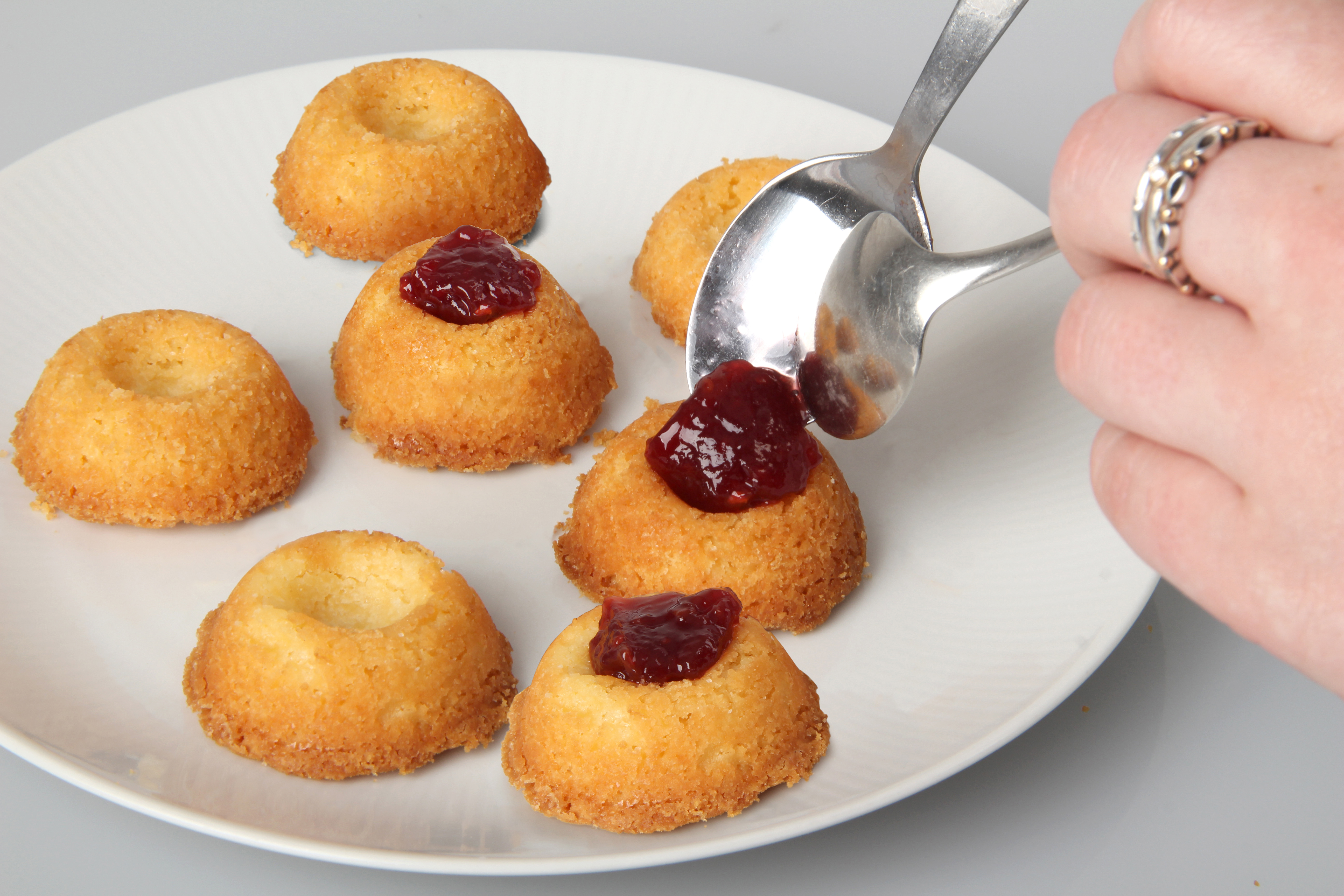 Teller mit kleinen, runden, goldenen Kuchen. Eine Hand fügt einem Kuchen mit zwei Löffeln Marmelade hinzu.
