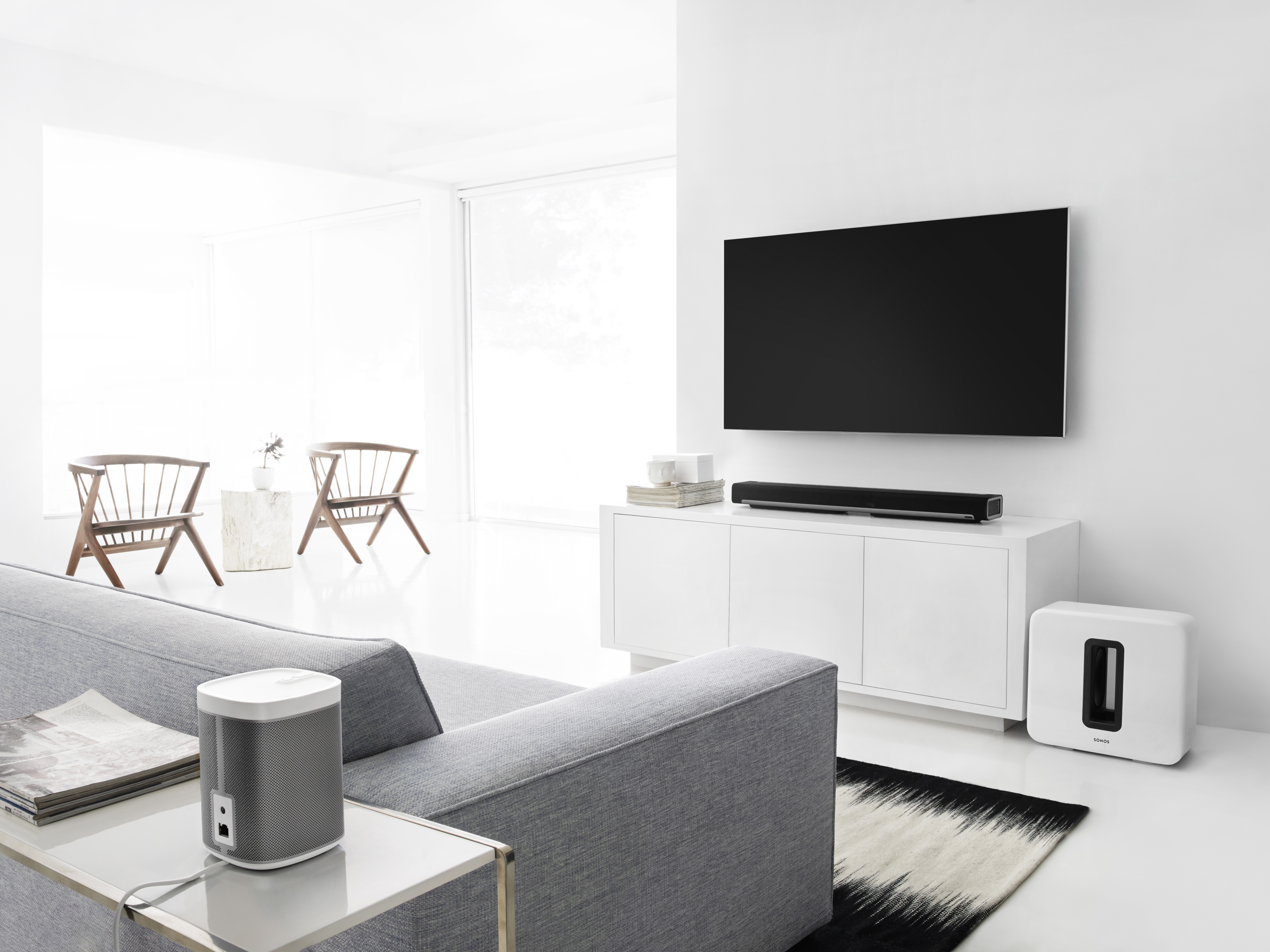 Ein modernes Wohnzimmer mit grauem Sofa, weißen Möbeln, einem Fernseher und einem Sonos-Lautsprecher.