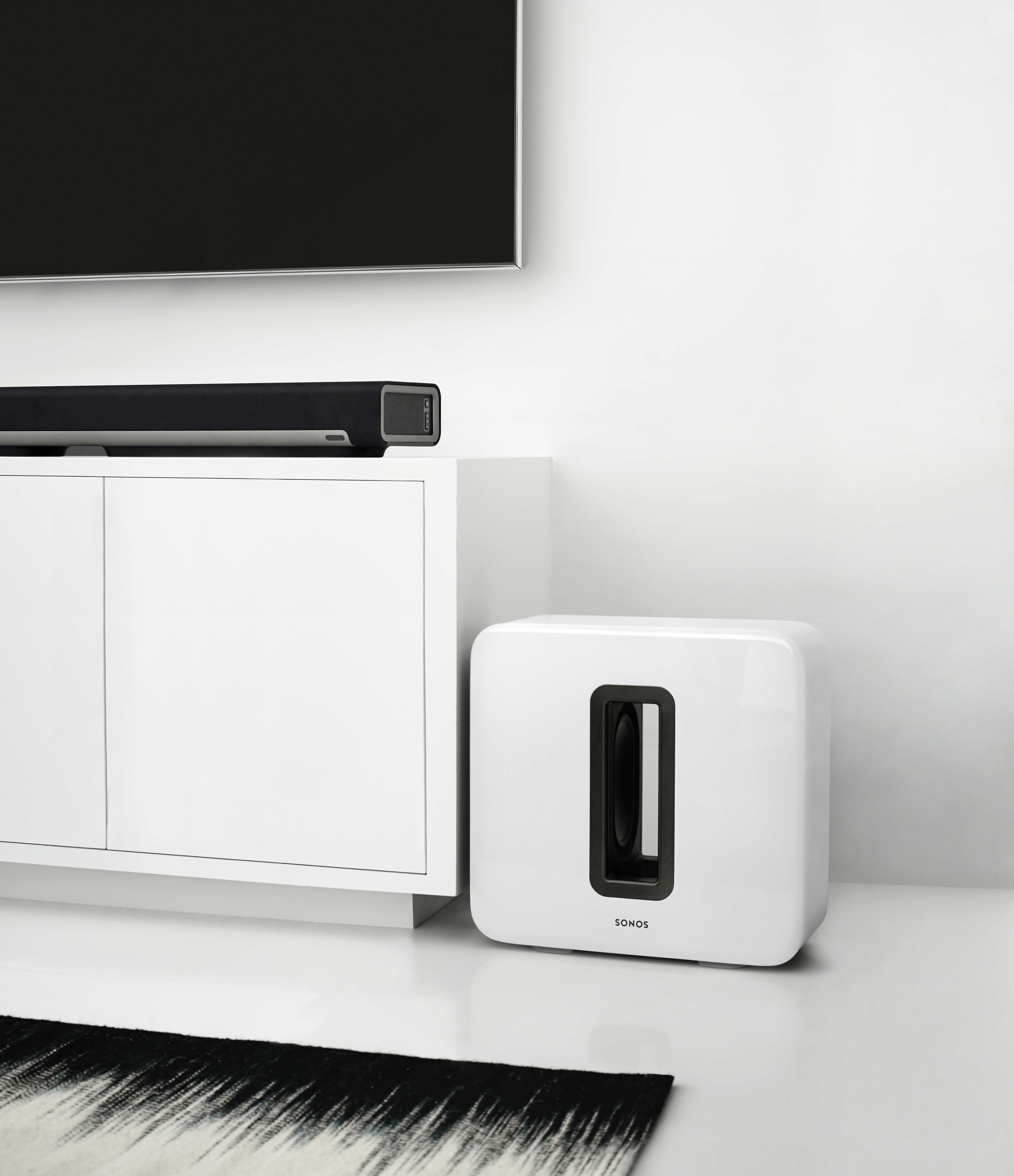 Weißer Subwoofer und Soundbar auf weißem Schrank. Schwarzer Teppich auf dem Boden. Fernseher an der Wand.