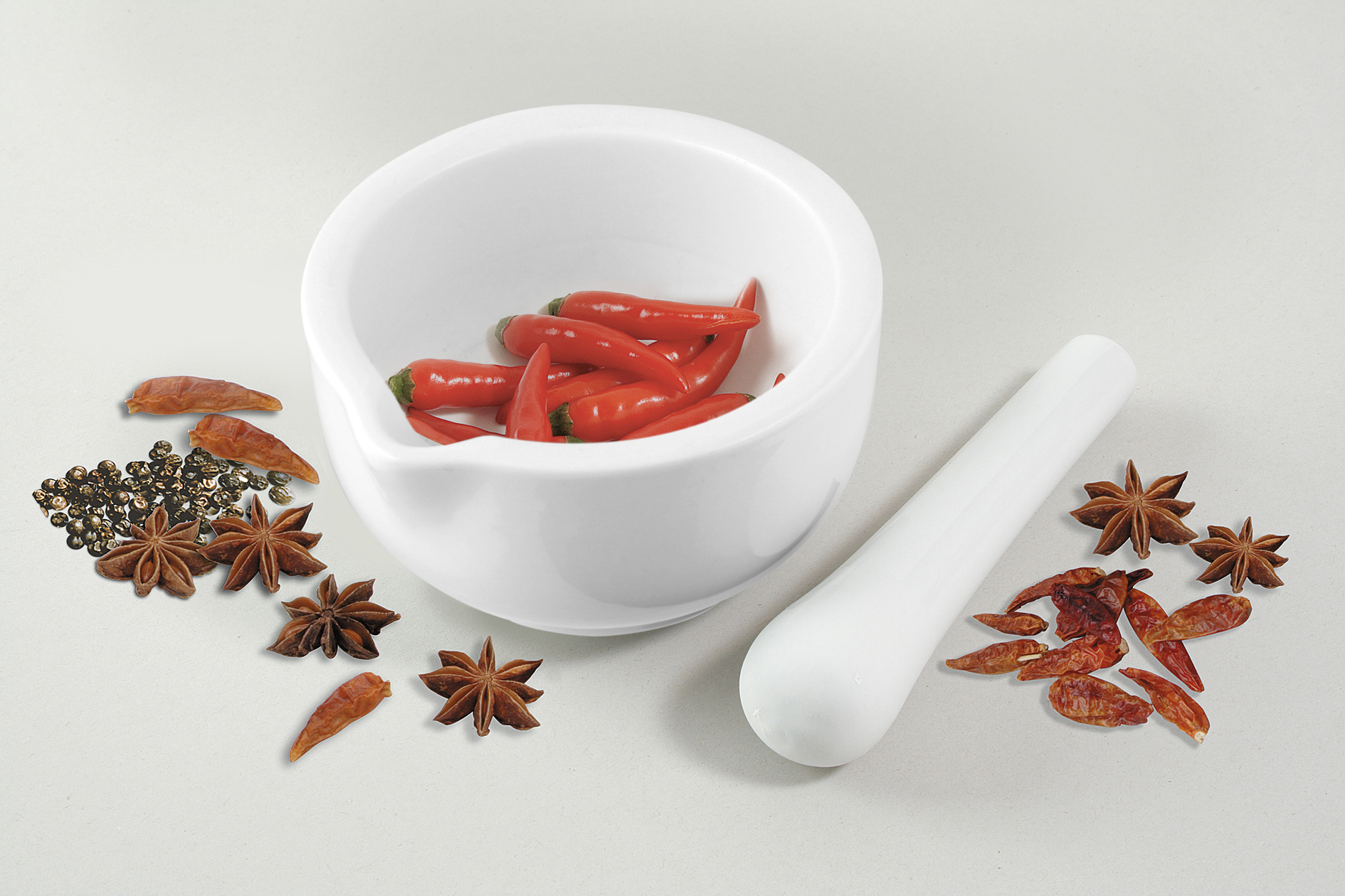 Weiße Schüssel mit roten Chilischoten. Sternanis, Pfefferkörner und getrocknete Chilis sind daneben.