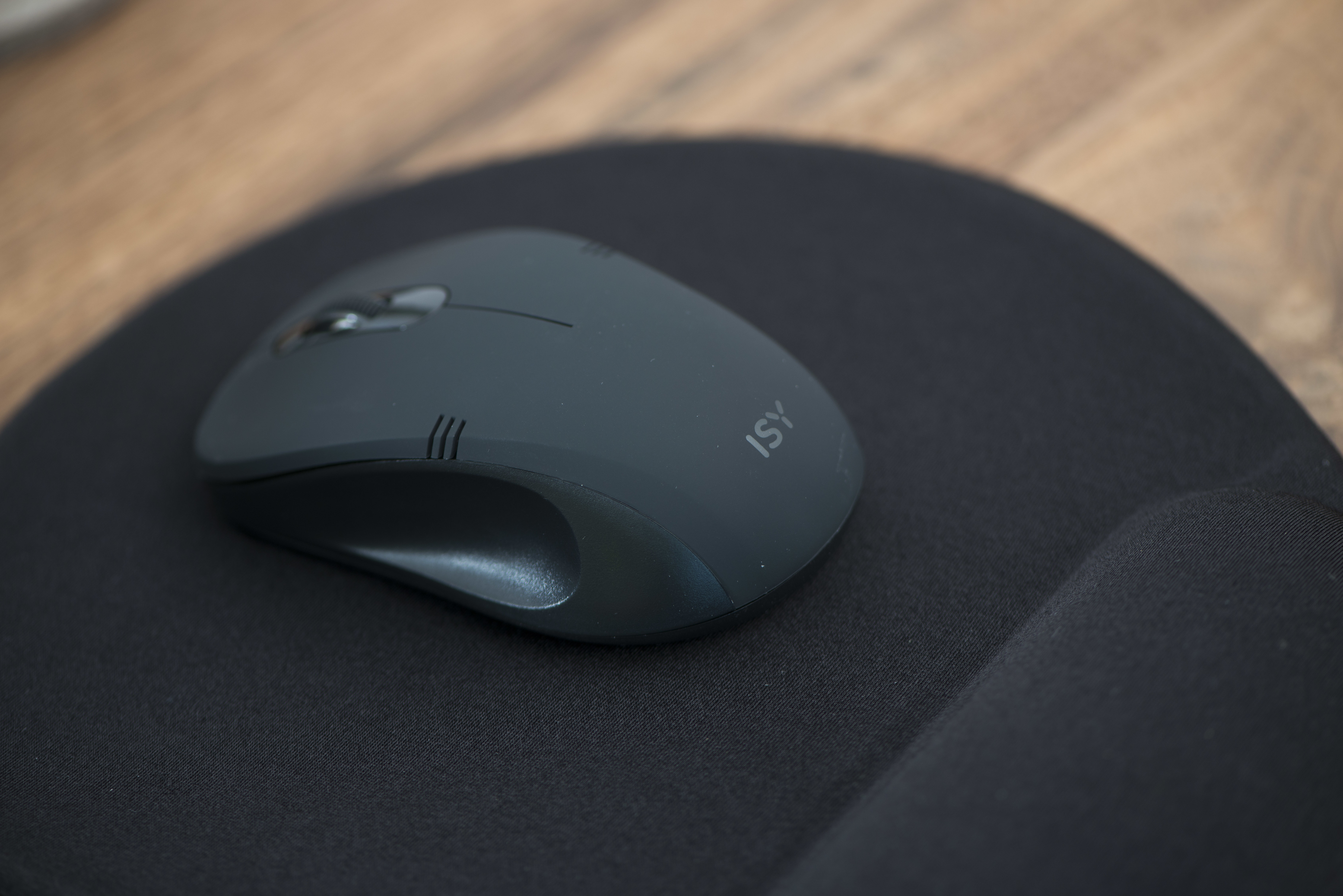 Une souris gris foncé sur un tapis de souris noir avec repose-poignet. La souris est de marque ISY.