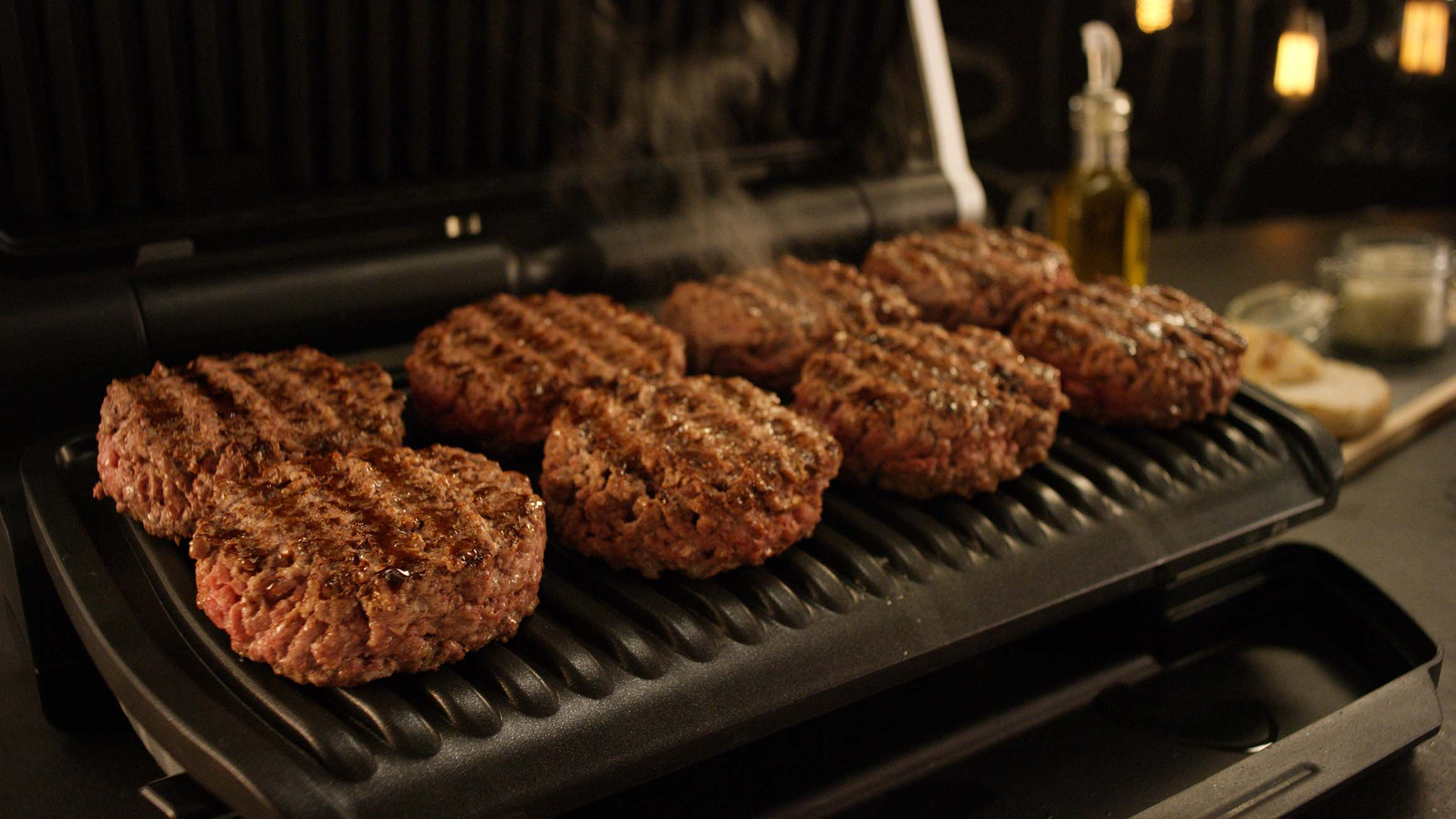 Gegrilde hamburgerpasteitjes op een grill, met stoom die opstijgt en andere items op de achtergrond.