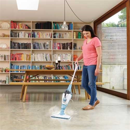 Woman using a steam mop on the floor. Background: books and a table.