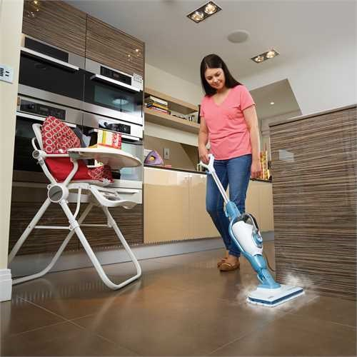 A woman using a steam mop in a kitchen, with a baby chair.