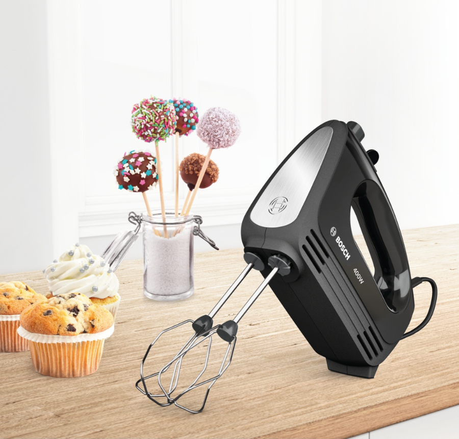 Zwarte handmixer op een houten tafel, met cupcakes en cakepops op de achtergrond. Witte achtergrond.