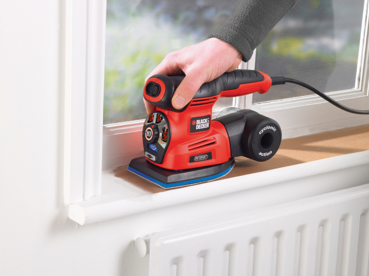 Fekete-piros Black & Decker csiszoló. Egy kéz működteti egy fehér ablakkereten.