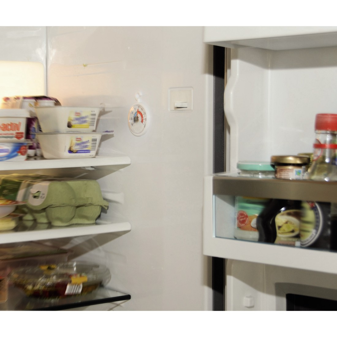 Open refrigerator interior with shelves holding food items like eggs, butter, and jars. A thermometer is on the wall.