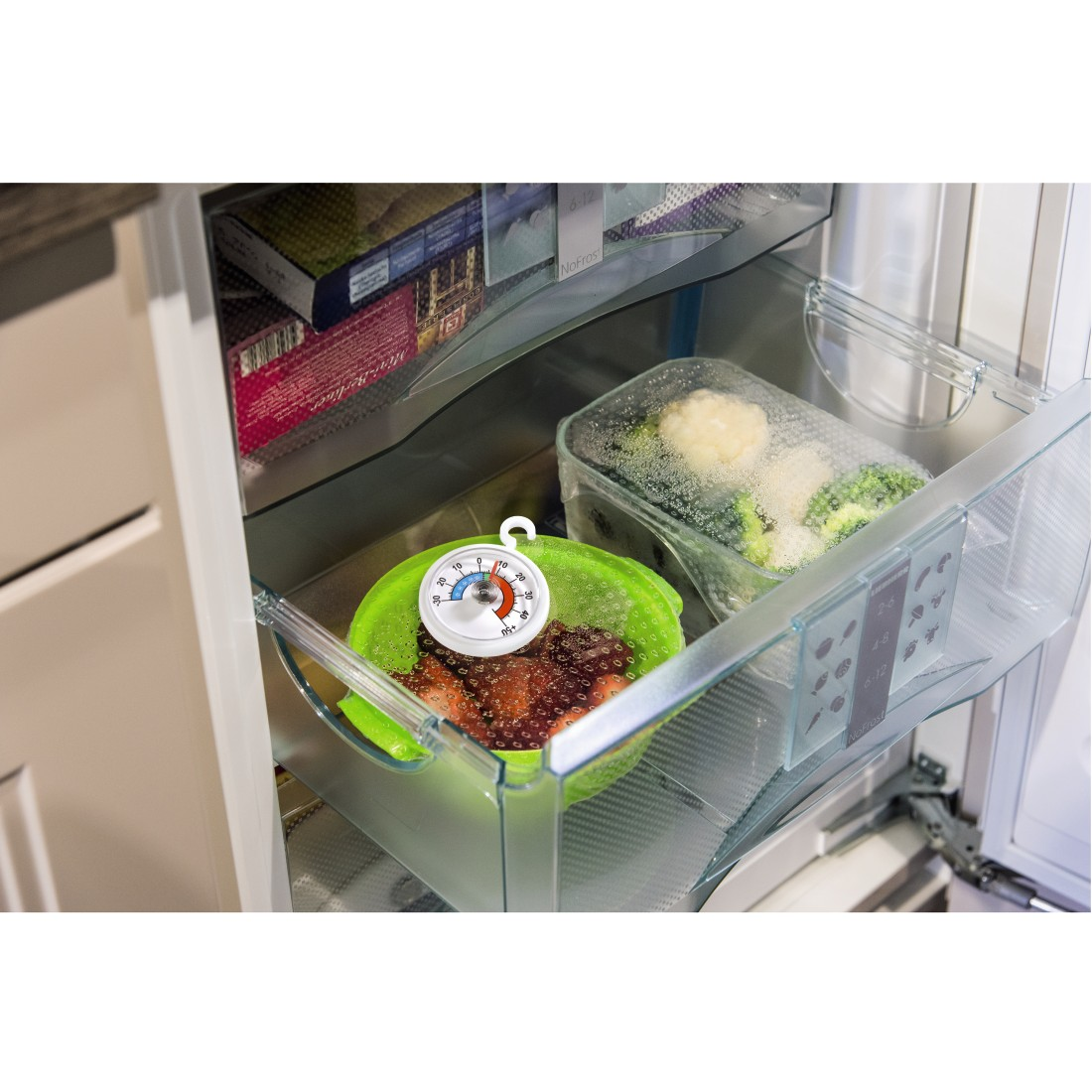 Inside a fridge: A thermometer hangs over a green bowl of strawberries. Broccoli and cauliflower are in a clear container.