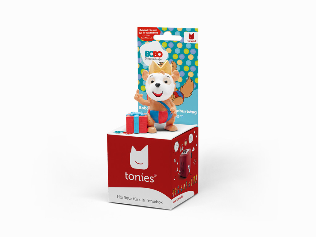 Eine Toniebox-Figur, Bobo, steht auf einer rot-weißen Box. Ein Geschenk steht neben ihm.