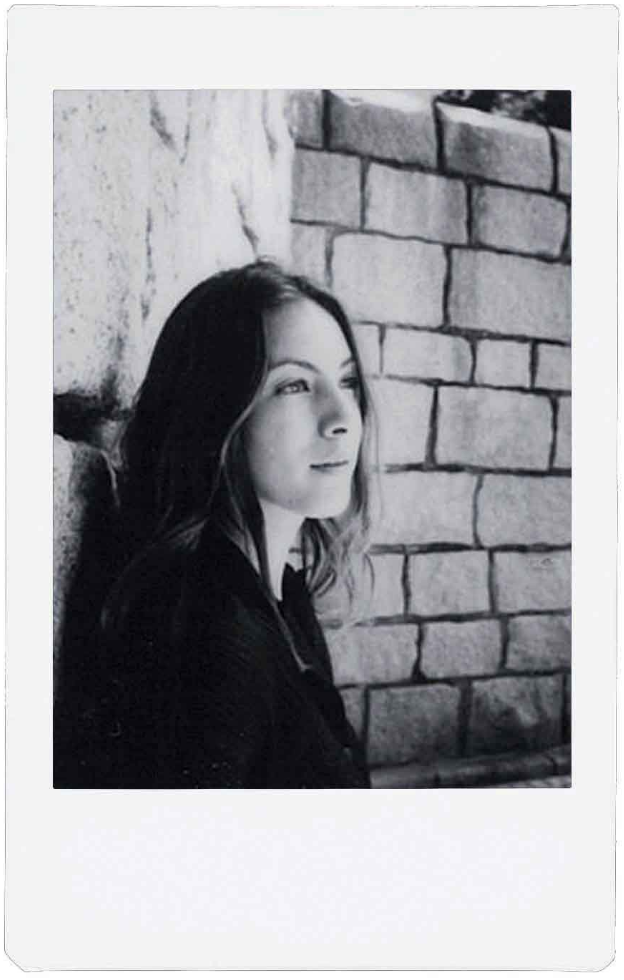 A black and white photo, a woman leans against a brick wall, looking to the side.