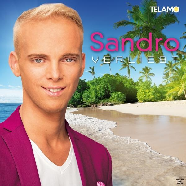 Mann mit blonden Haaren, lächelt, trägt rosa Blazer, Strandhintergrund.