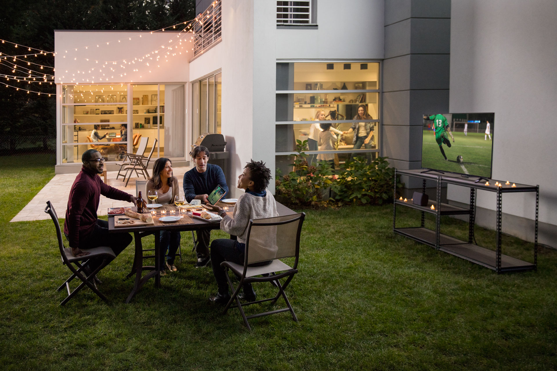 People sitting around a table outdoors. A TV shows a football game in the background.