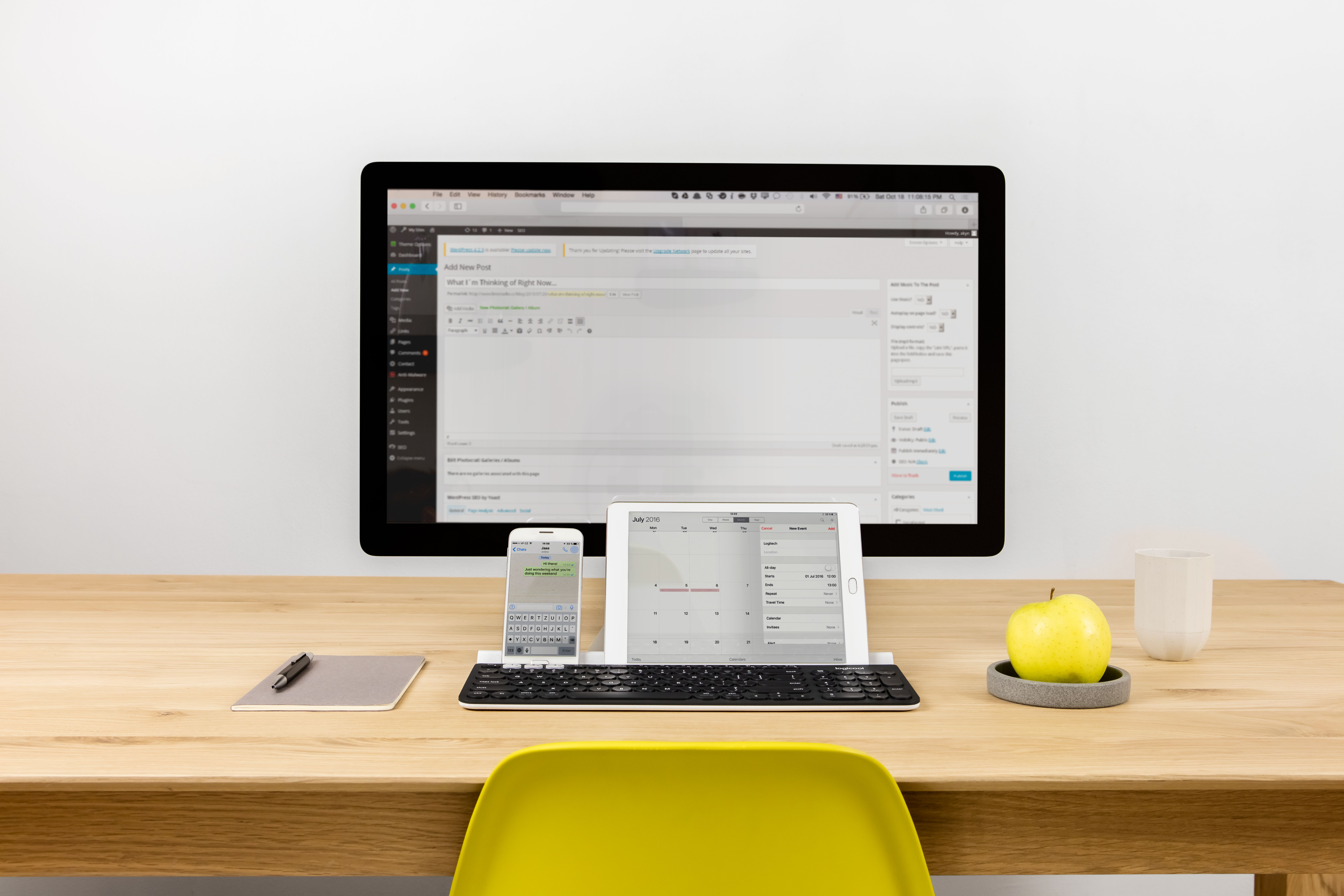 Bureau-opstelling met computer, tablet, telefoon, toetsenbord en appel. Helder licht op witte muur.