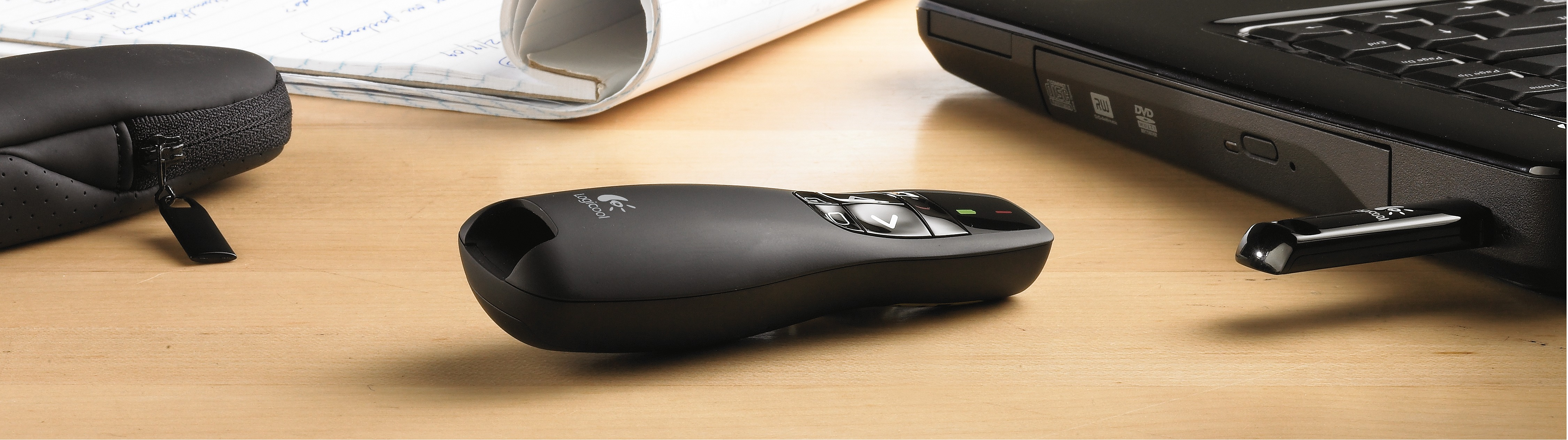 Zwarte Logitech-presenter met USB-ontvanger aangesloten op een laptop. Een pen en notitieboek liggen op het houten bureau.