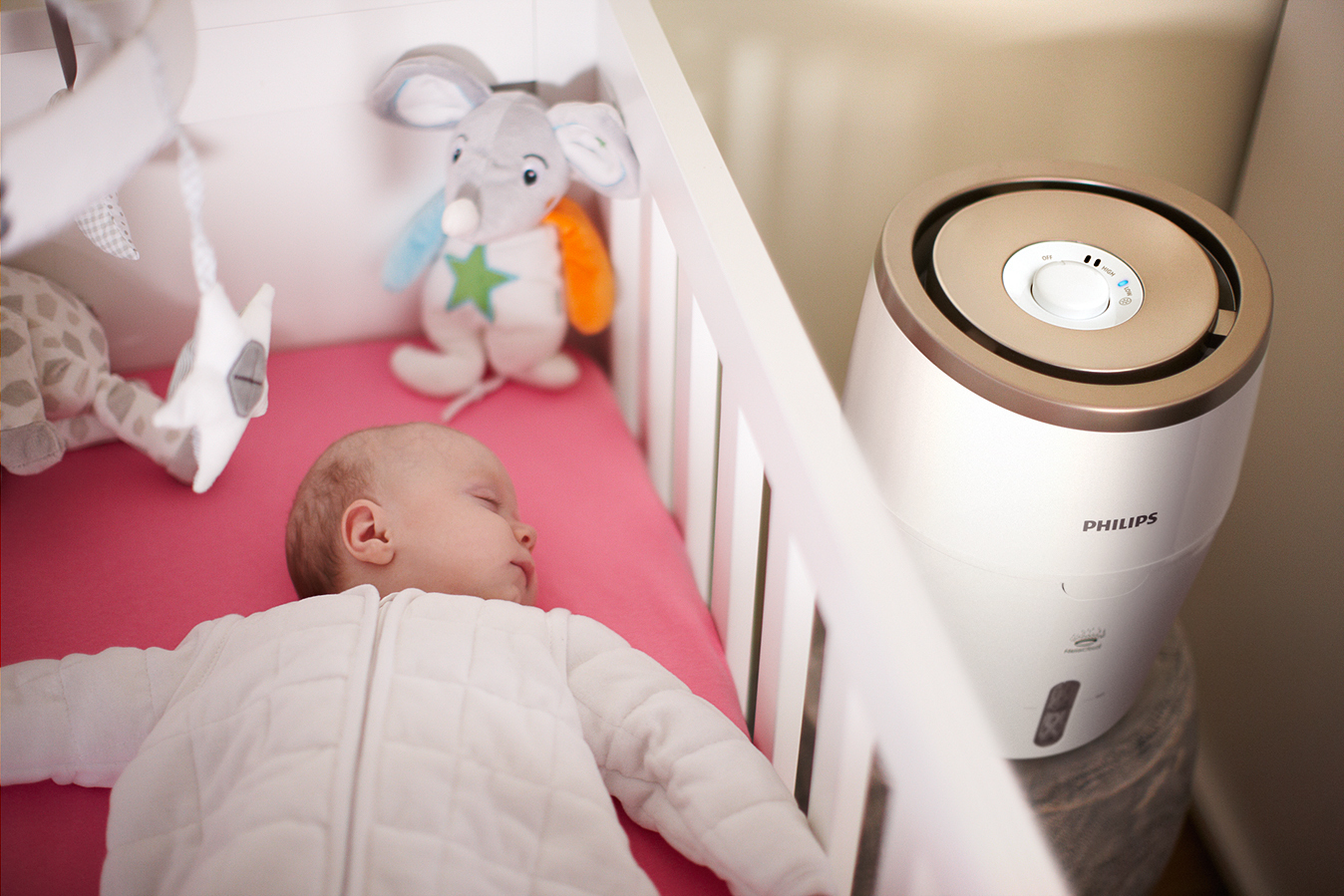 Ein Baby schläft in einem weißen Kinderbett neben einem Philips Luftbefeuchter. Ein Stofftier und Mobile sind über dem Bett.