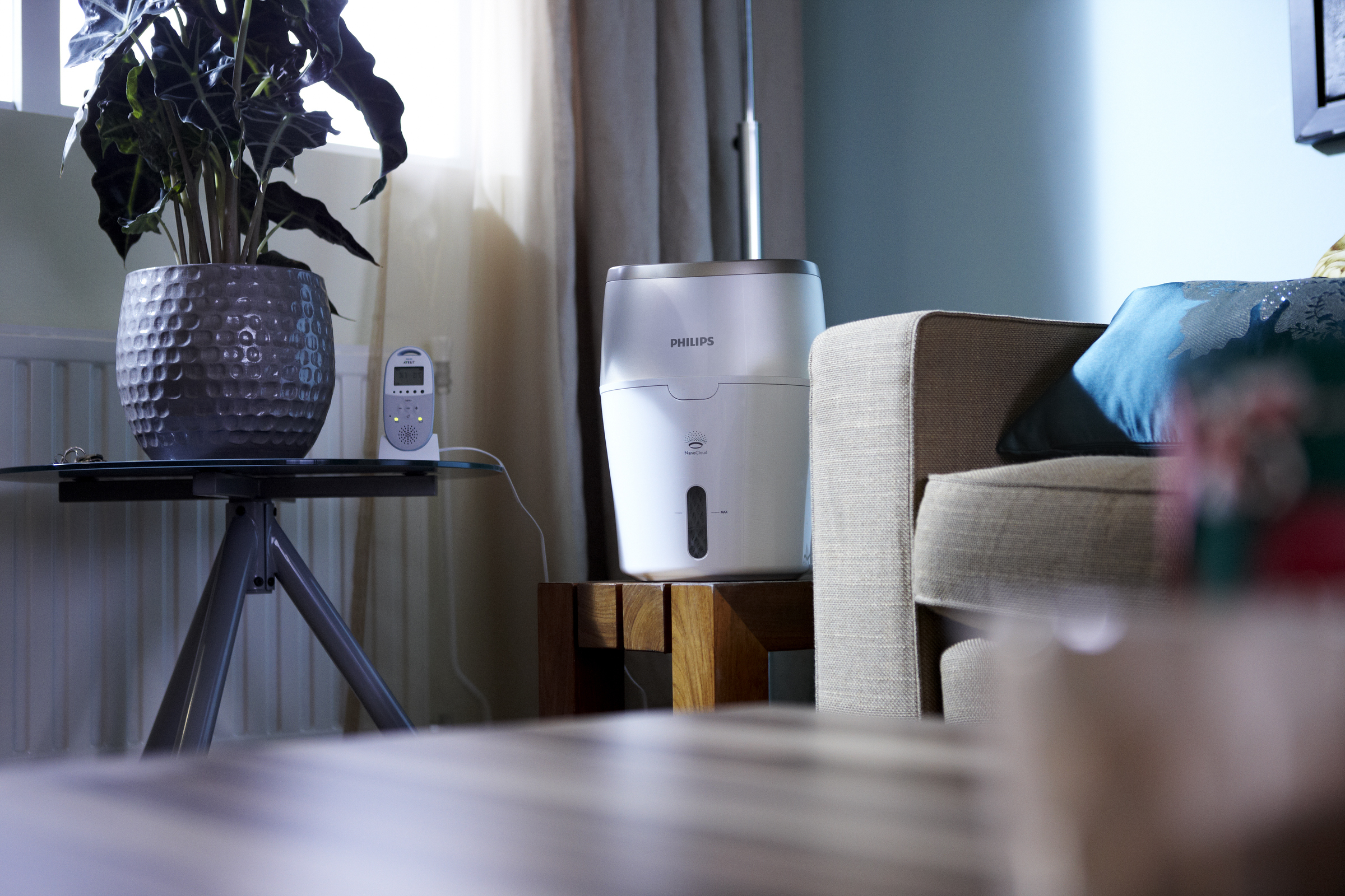Moderner Luftbefeuchter auf einem Holztisch mit Pflanze und Babyphone, in einem Wohnzimmer.
