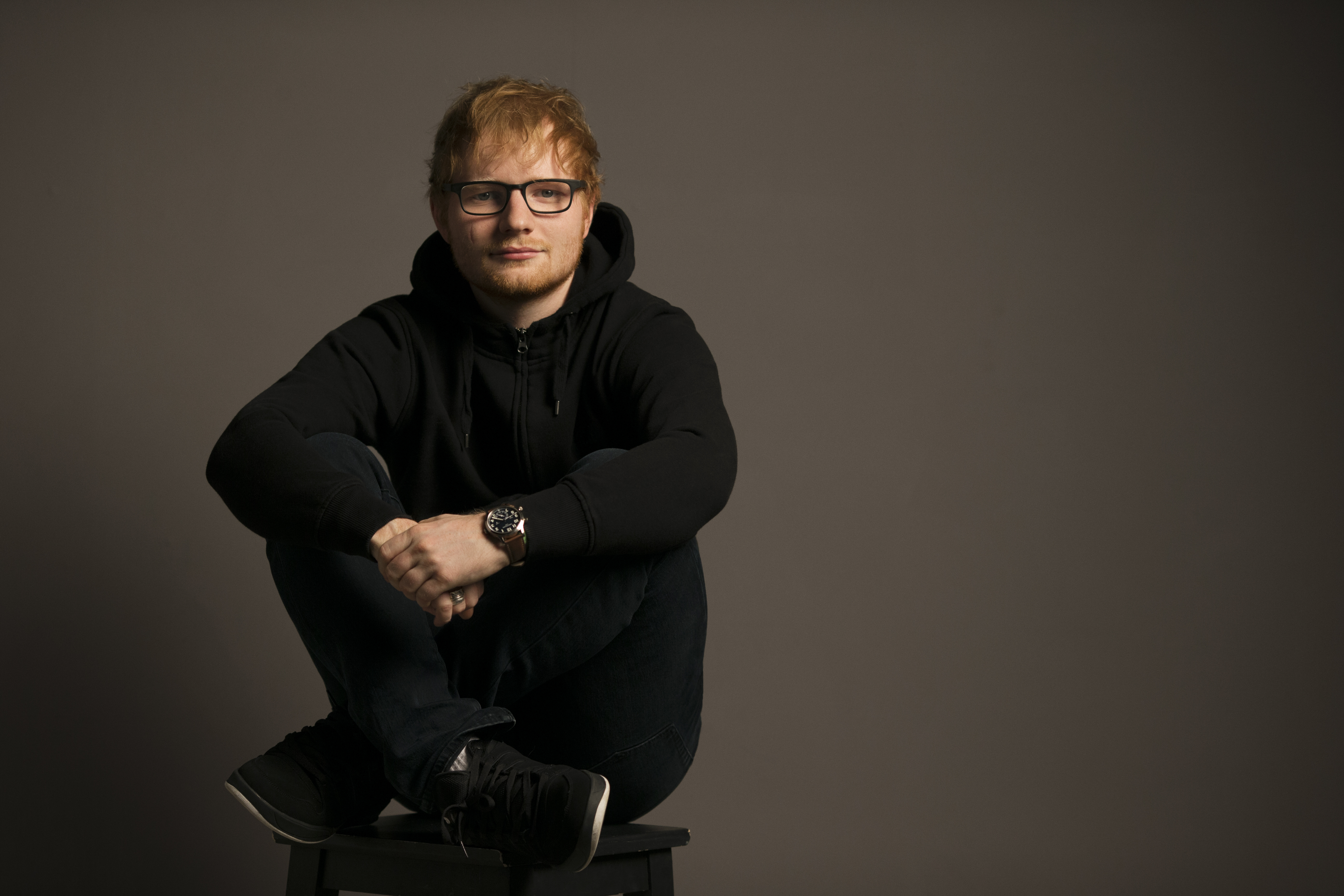 Ed Sheeran sitzt auf einem Hocker, trägt eine Brille und einen schwarzen Kapuzenpullover.