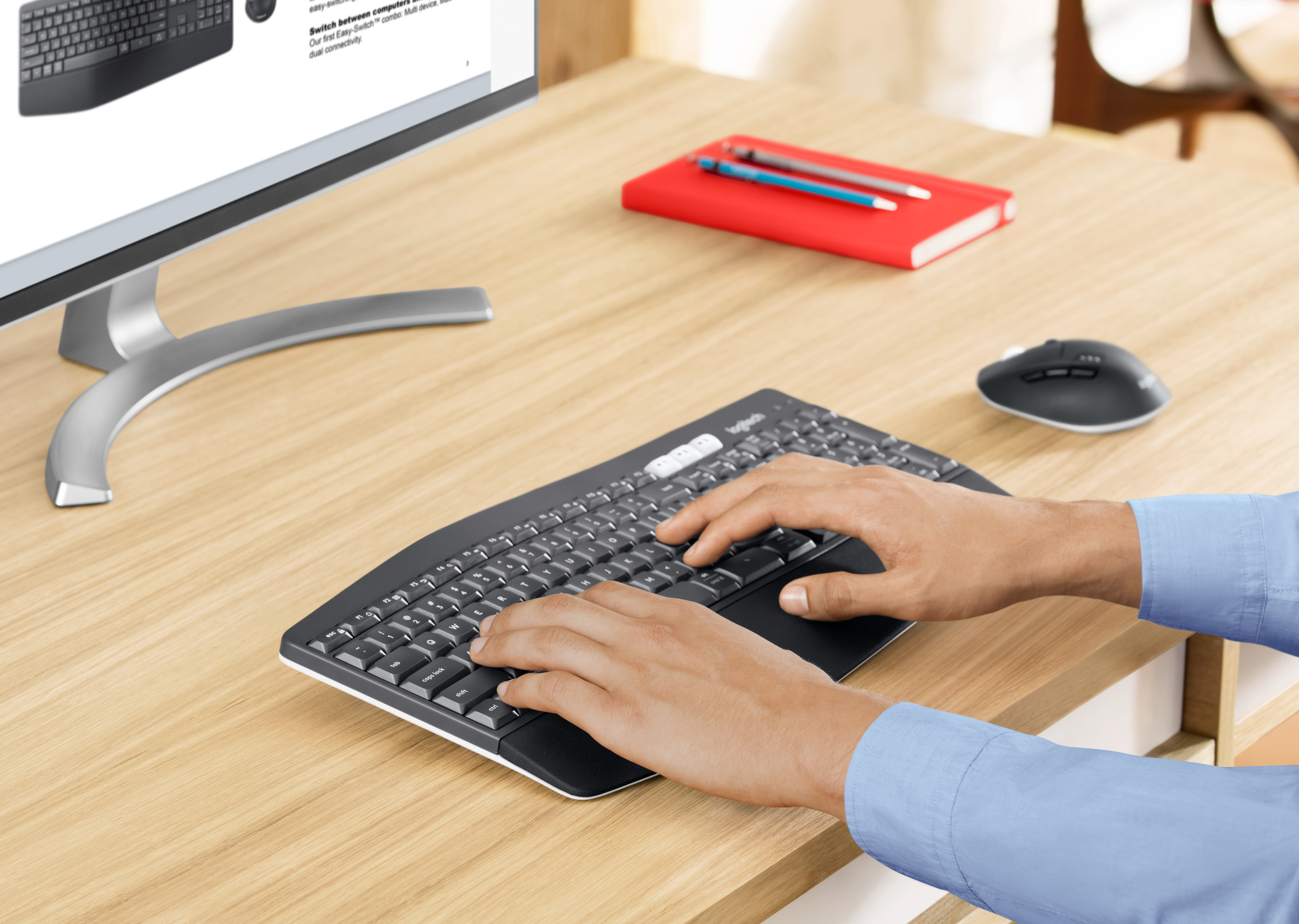 Hands on a curved keyboard. Monitor, red notebook, and mouse on a wood desk.