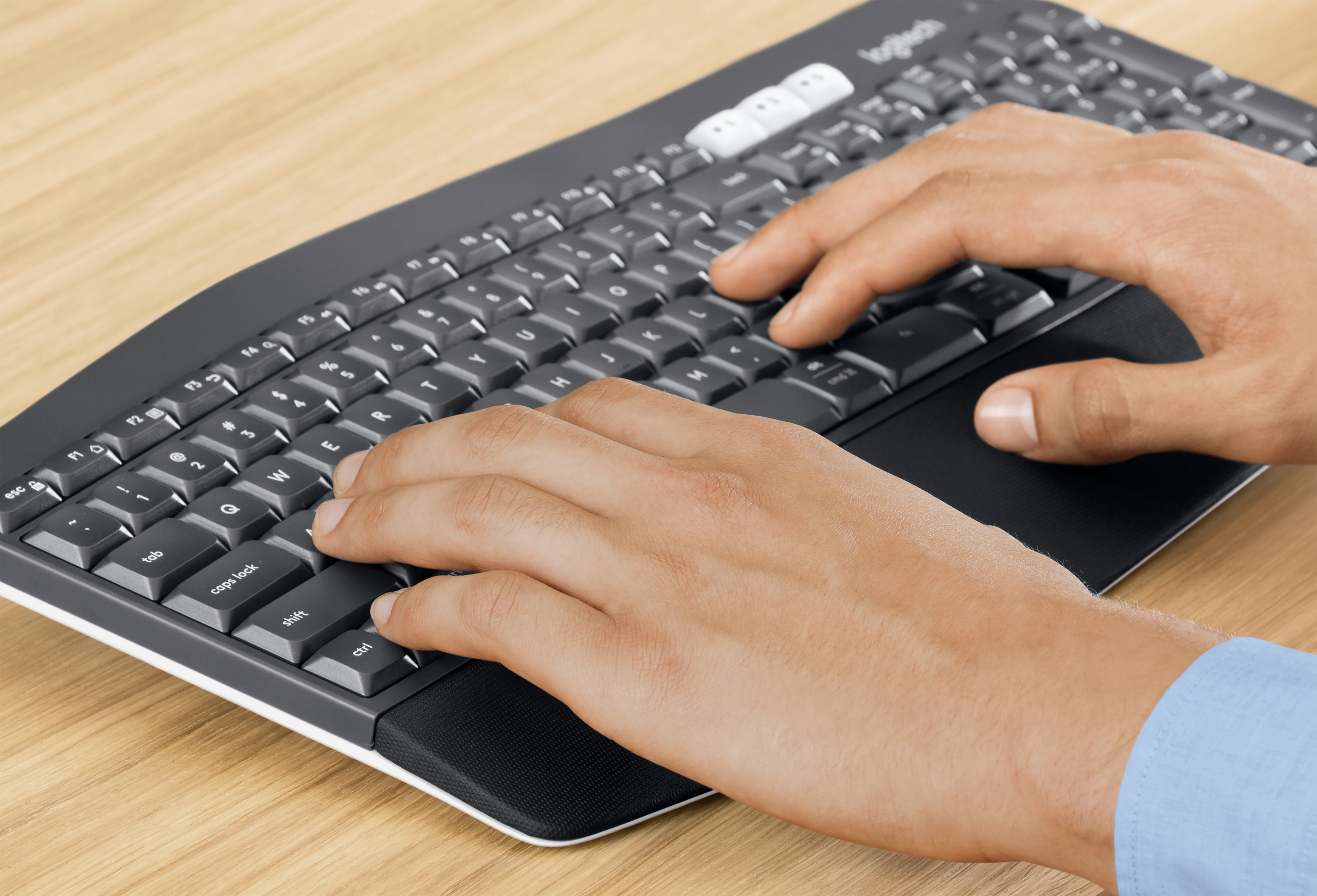 Hands typing on a black Logitech keyboard resting on a wooden surface.