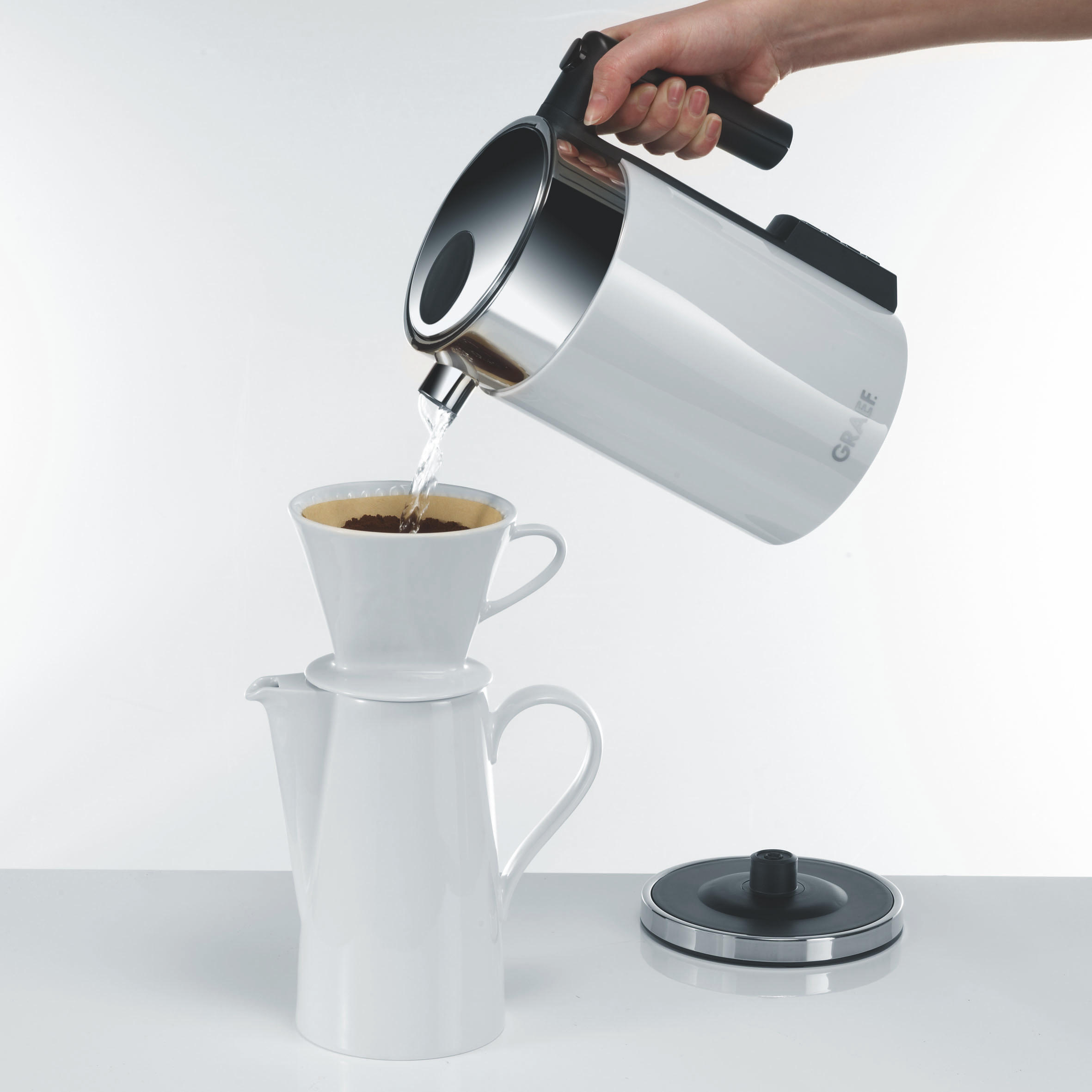 Eine Person gießt Wasser aus einem weißen Wasserkocher in einen Kaffeefilter.