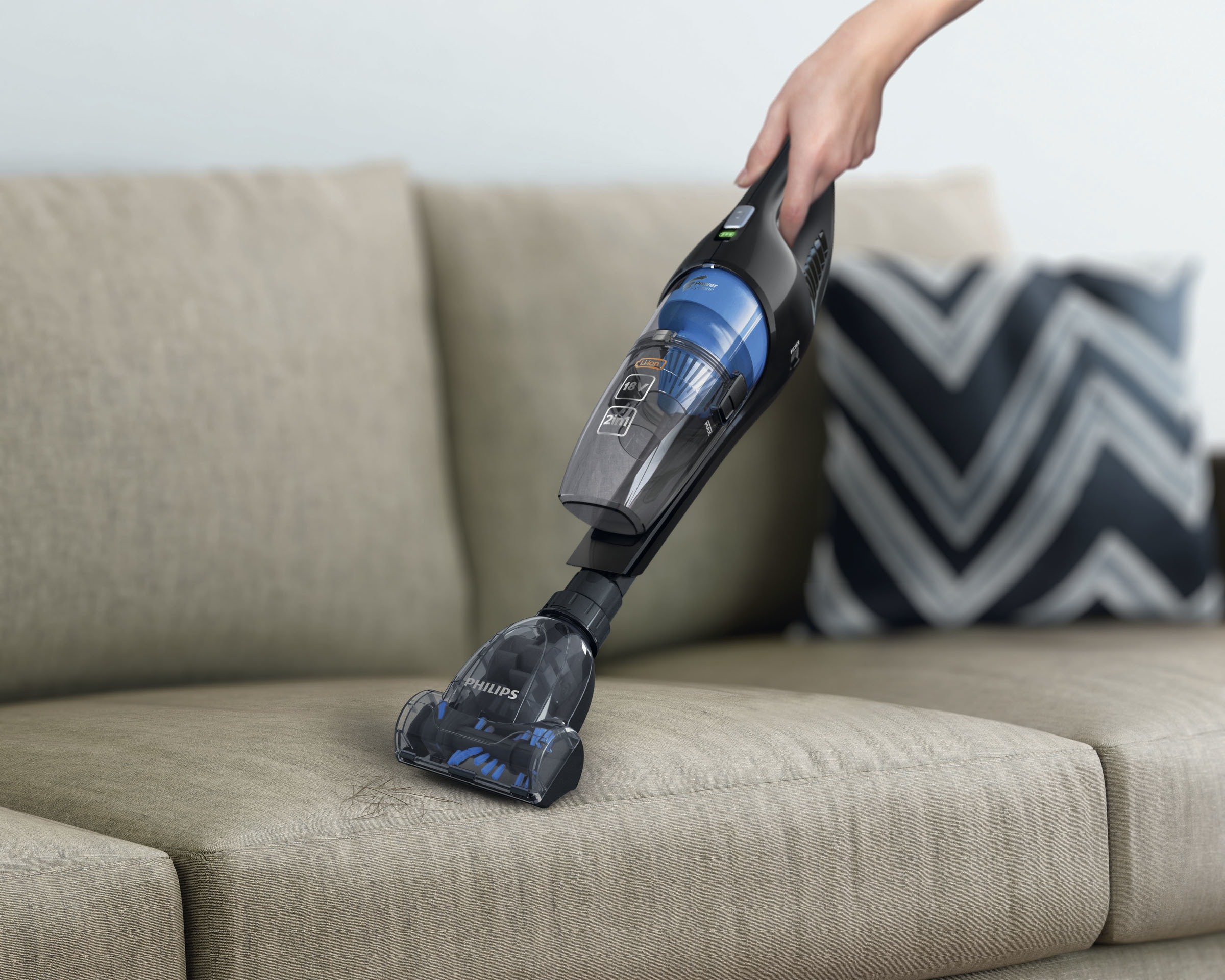 Ein Handstaubsauger saugt einen beigen Sofa ab. Der Staubsauger ist schwarz und blau. Ein Kissen mit weißem und schwarzem Muster liegt dahinter.
