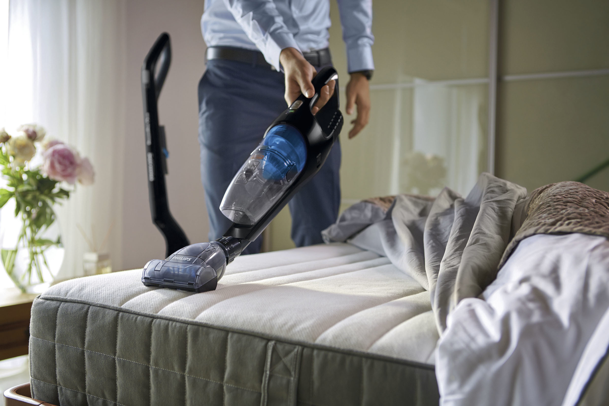 Ein Mann saugt ein Bett mit einem Philips Staubsauger. Ein Blumenstrauß ist im Hintergrund zu sehen.