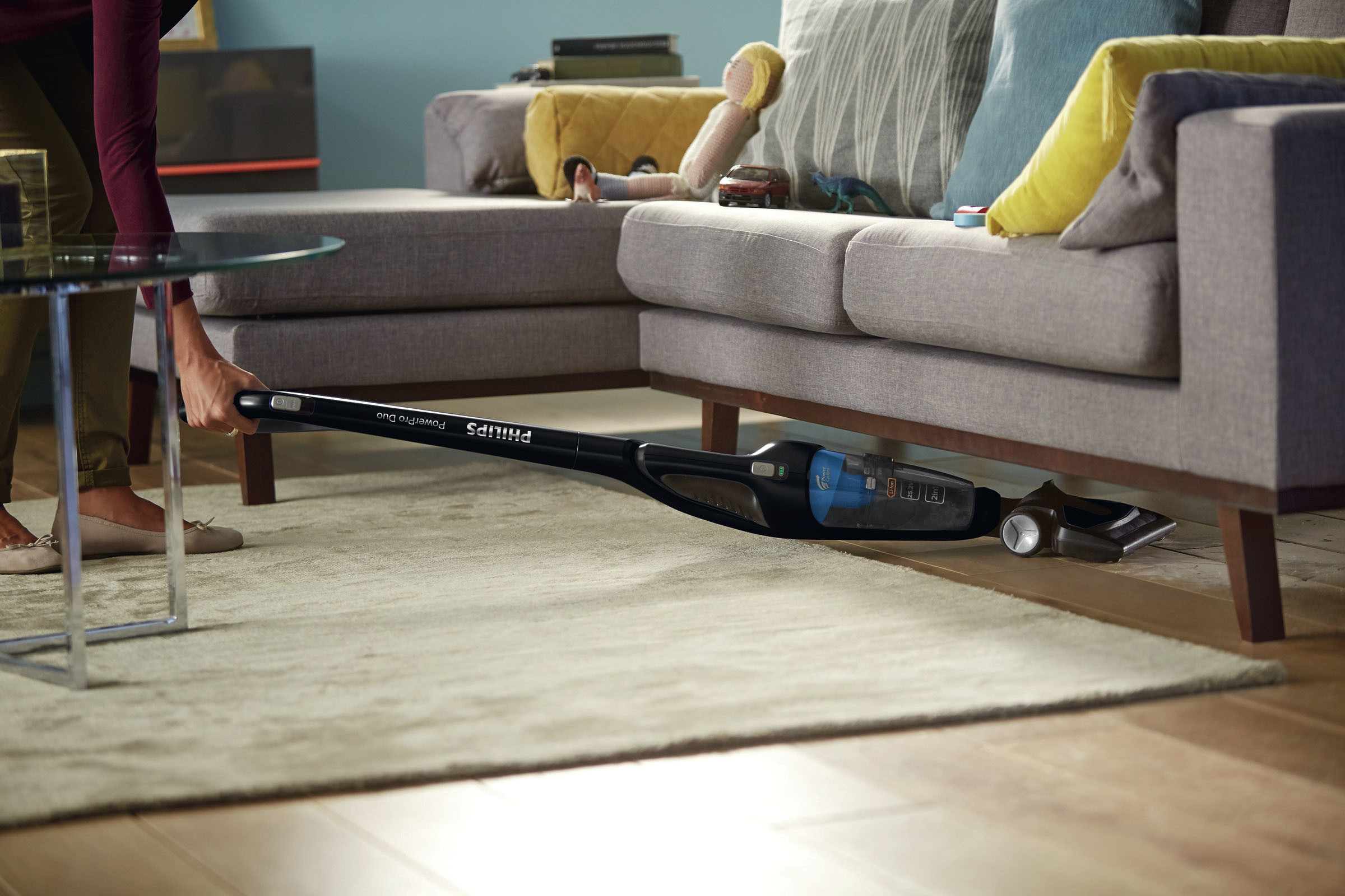 Person saugt mit einem schwarzen und blauen Philips Staubsauger unter einem grauen Sofa auf einem beigen Teppich.