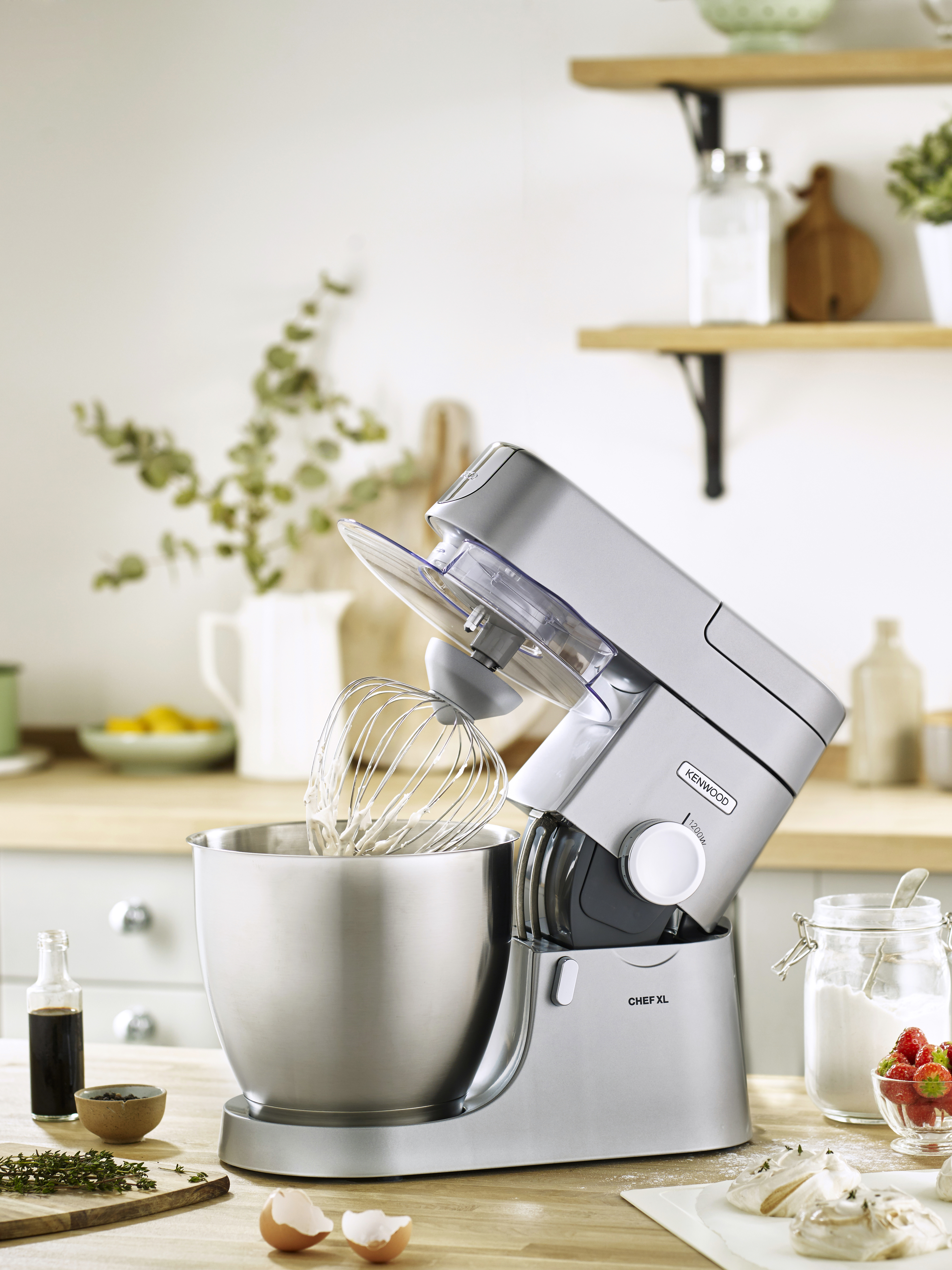 Ein silberner Küchenmixer mit Schneebesenaufsatz steht auf einer Holztheke. Der Hintergrund zeigt Küchenutensilien.
