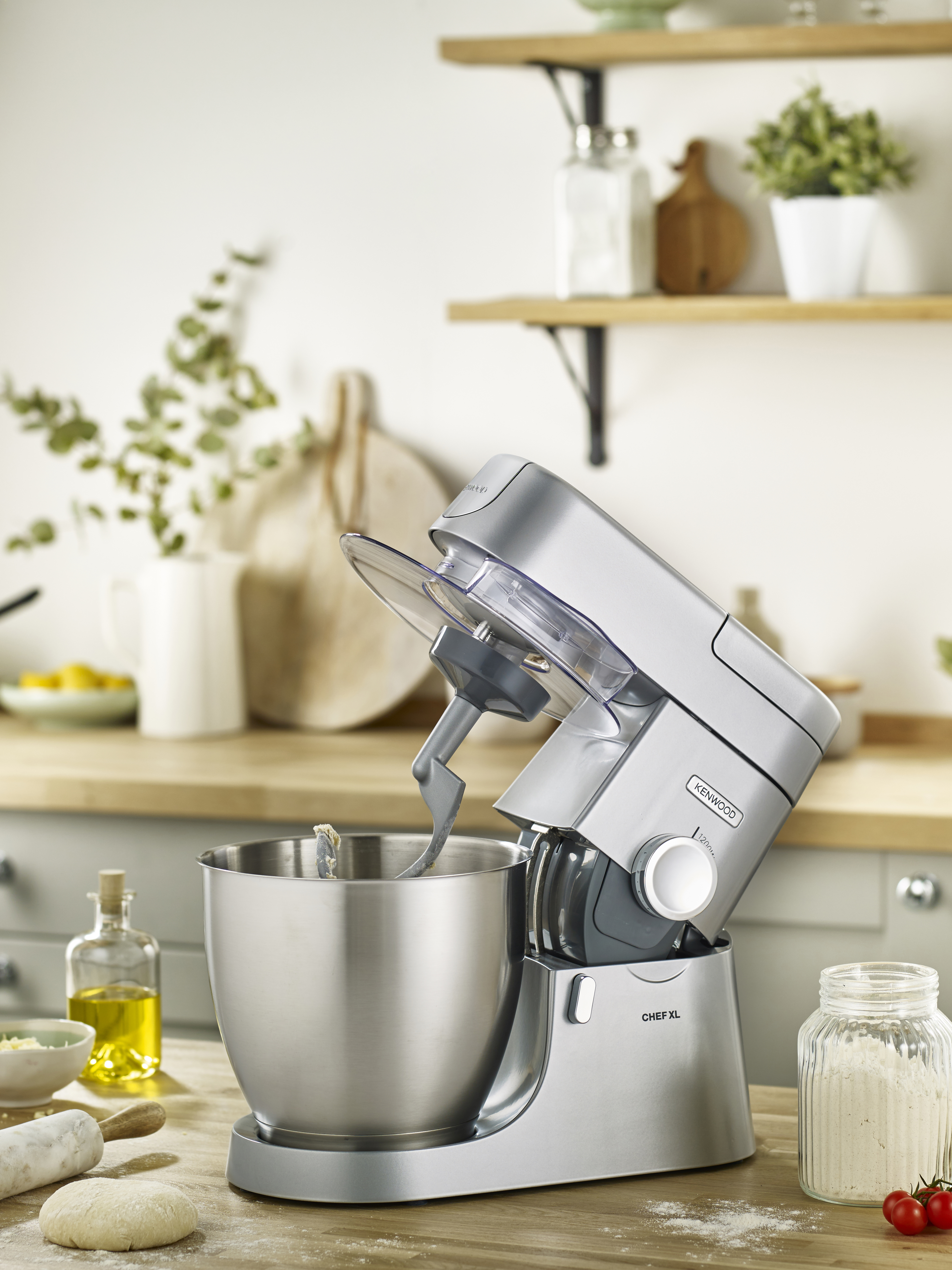 Silberner Küchenmixer mit Schüssel, Mehl und Ölflaschen auf einer Holz-Arbeitsplatte.