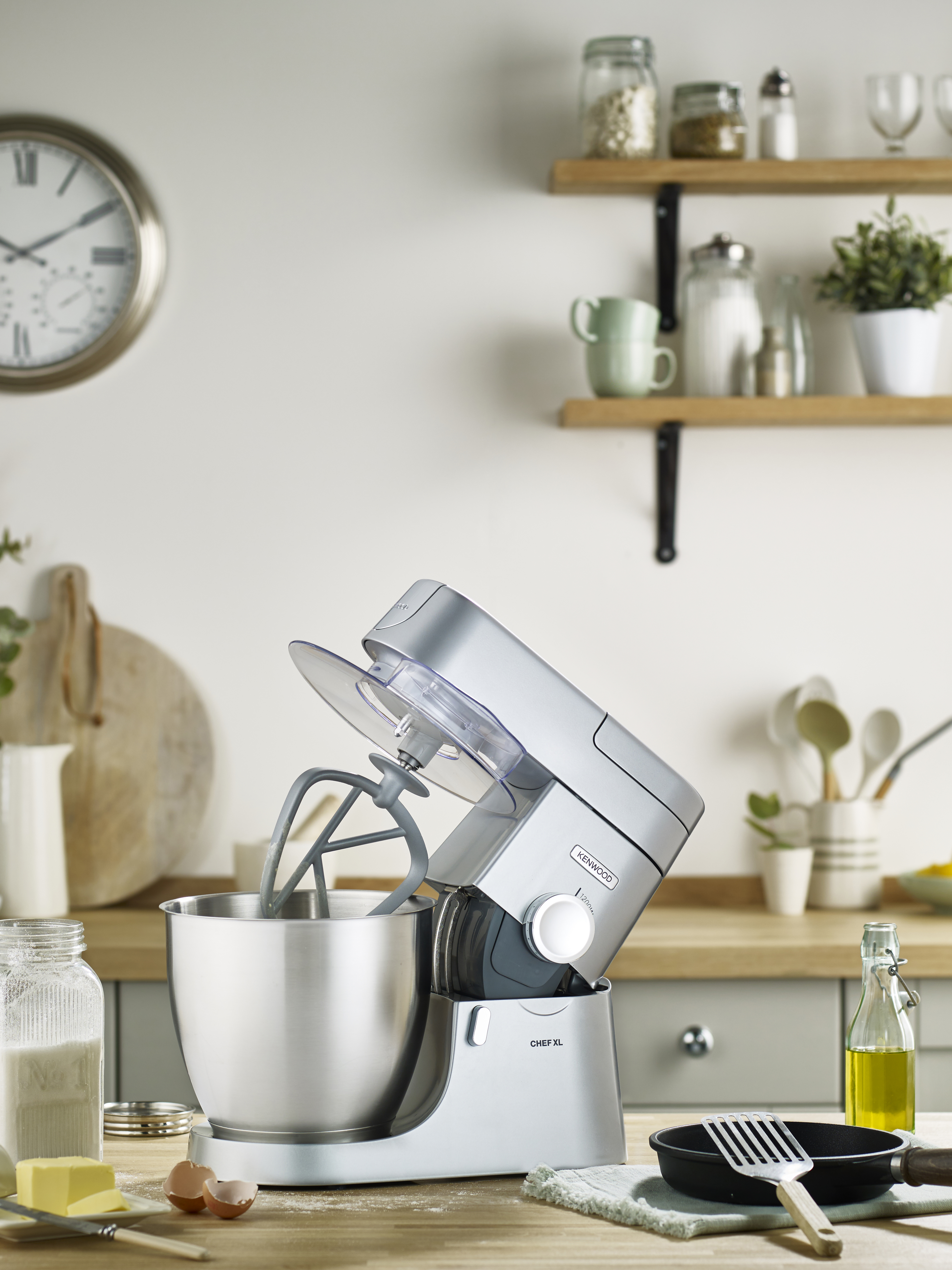 Ein silberner Küchenmixer mit Schüssel auf einer Holztheke. Zutaten und Uhr im Hintergrund.
