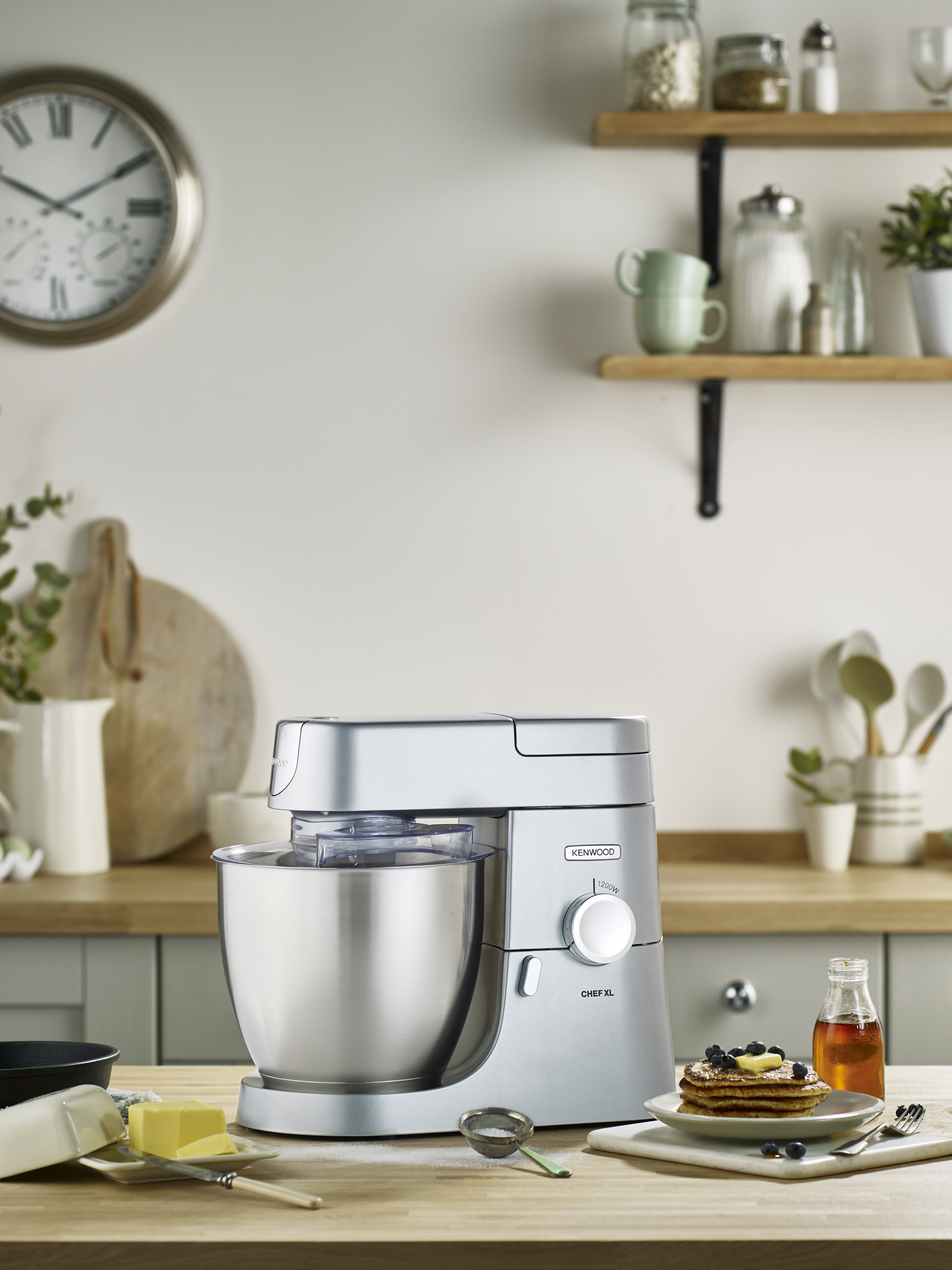 Küchenszene mit einem Kenwood Chef XL Mixer. Tisch mit Pfannkuchen, Butter und Sirup. Uhr und Pflanzen.