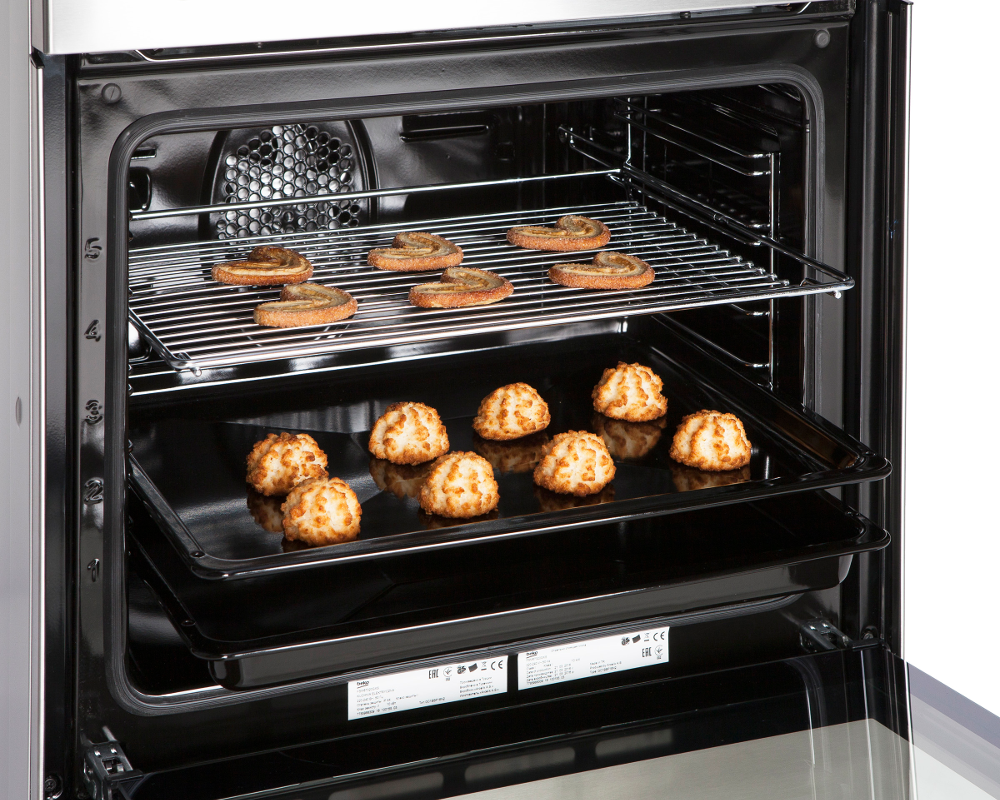 In een oven, twee roosters met gebakken goederen. Bovenste rooster heeft gebak. Onderste rooster heeft kokosmacarons op een bakplaat.