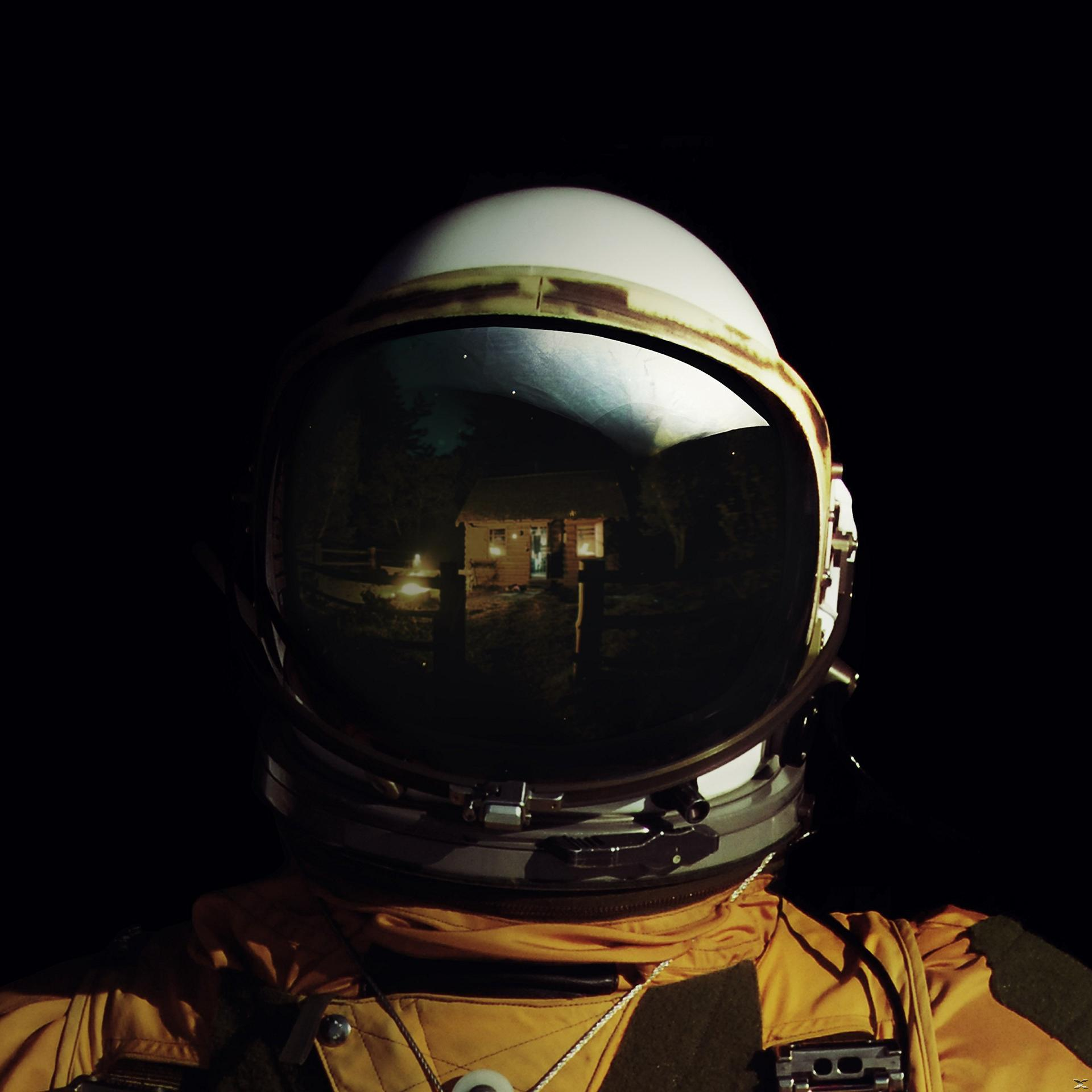 Astronautenhelm, der eine Hütte im Wald reflektiert. Der Hintergrund ist dunkel. Der Astronaut trägt einen gelben Anzug.