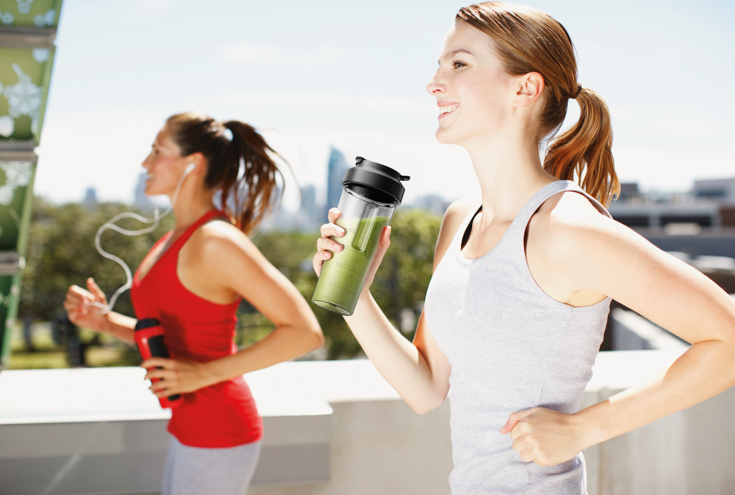 Twee vrouwen rennen buiten, een houdt een smoothiefles vast. Ze dragen sportkleding.