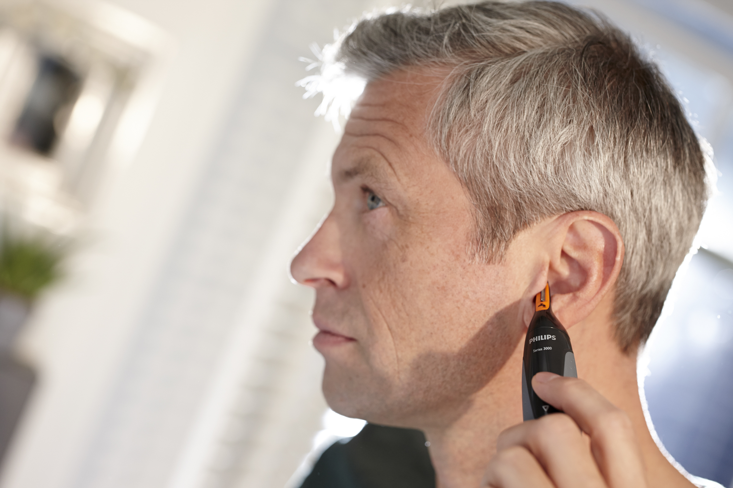 Mann trimmt Ohrenhaare mit einem kleinen schwarz-orangenen Trimmer. Seitenprofil.